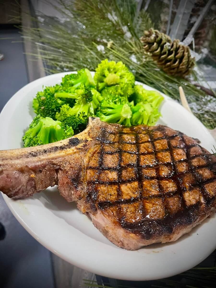 Grilled T-bone steak with crosshatch grill marks served with steamed broccoli on a white plate, with Christmas decorations including pine branches, pine cones, and a Christmas ornament in the background.
