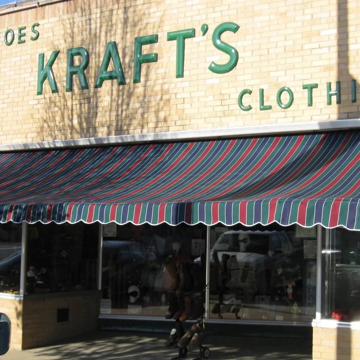 Exterior view of a clothing store named 'Kraft's Cloth' with a striped awning, glass windows, and a brick facade.