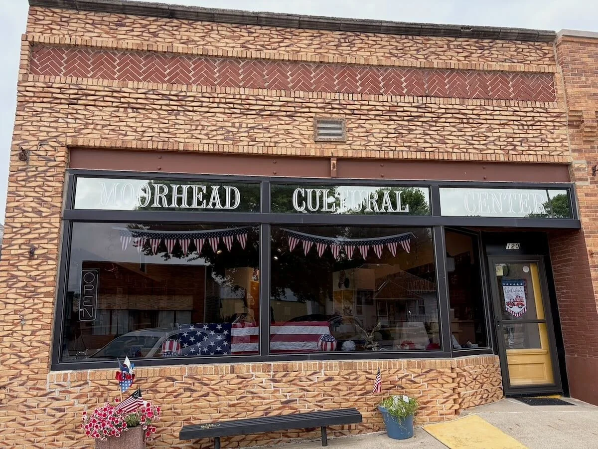 The exterior of the Morhead Cultural Center features a brick facade with large glass windows decorated with patriotic bunting and American flags. The entrance has a black door with patriotic decorations, and there are flower pots with flowers and small flags outside.