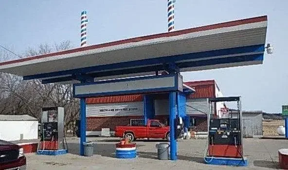 A vintage American gas station with a large canopy featuring red, white, and blue stripes, two fuel pumps, a red pickup truck, and a small building in the background.