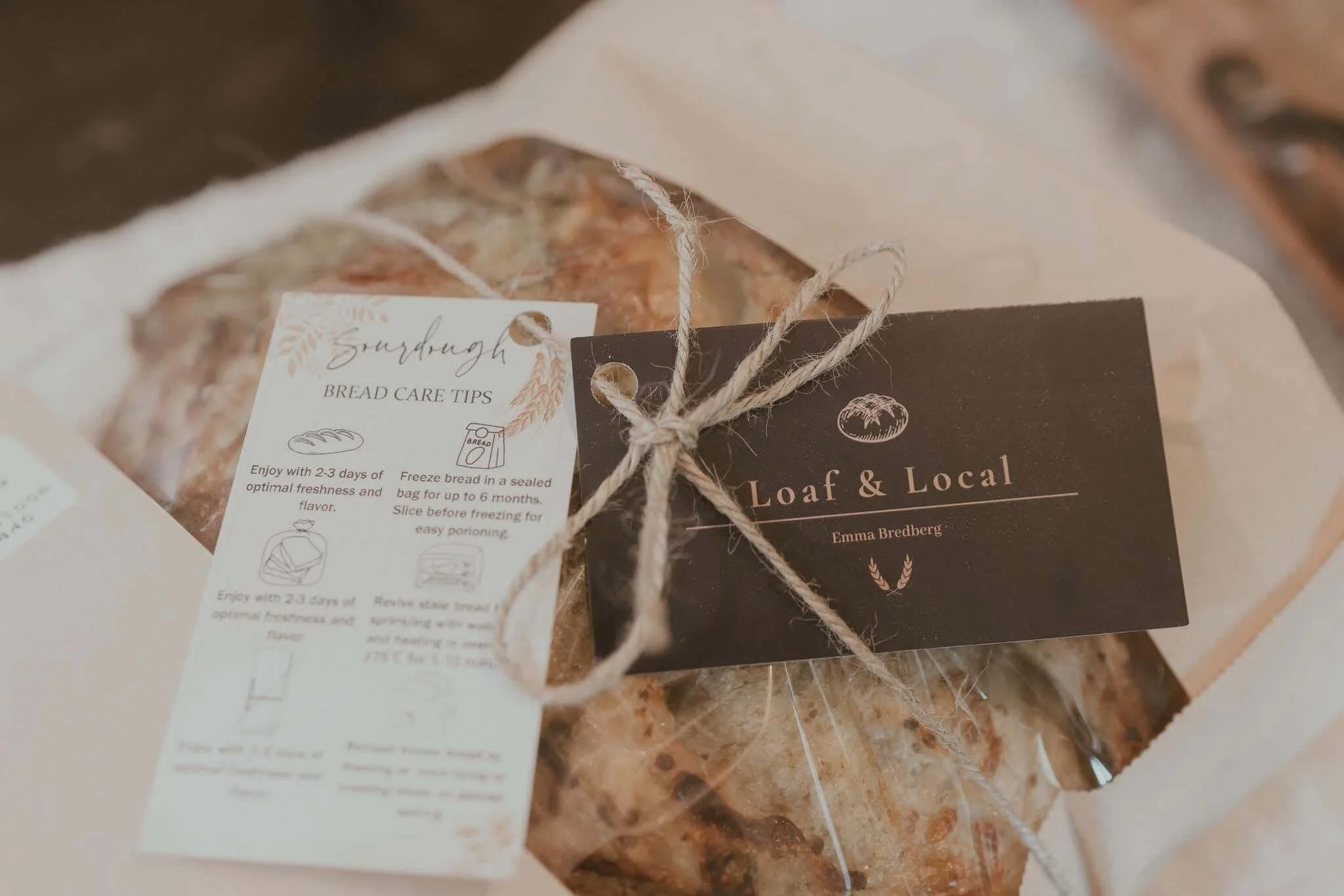 A packaged loaf of bread wrapped with twine, with a black and white label reading "Loaf & Local" and a smaller white tag with bread care tips and a sketch of bread.
