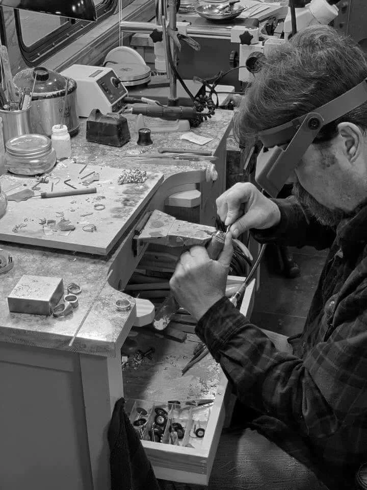 A person working on jewelry inside a jewelry workshop, using a soldering iron, with jewelry tools and equipment on the workbench.