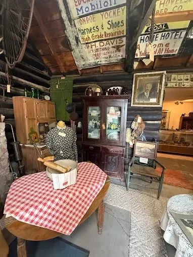 Vintage antique shop with a wooden cabinet, a table with a red-checked tablecloth, a mannequin with a polka-dot dress, framed pictures, and various signs on the walls.