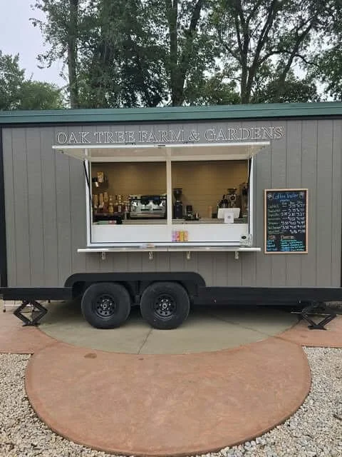 A mobile coffee stand named 'OAK TREE FARM & GARDENS' set up on a trailer with two tires. The stand has a serving window with a white counter, coffee equipment inside, and a chalkboard menu hanging on the right side. The stand is parked outdoors with trees in the background and a circular paved area in front.