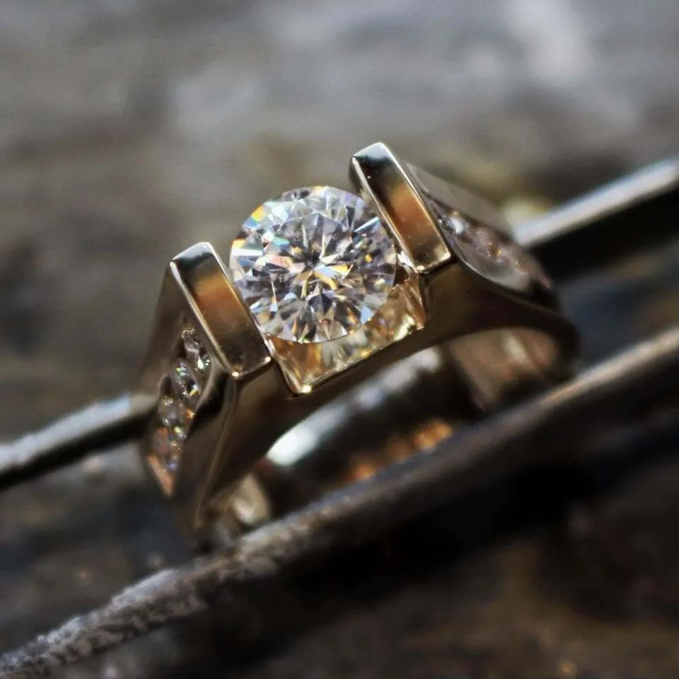 Close-up of a silver ring with a large central diamond and smaller diamonds on the band, resting on a textured surface.