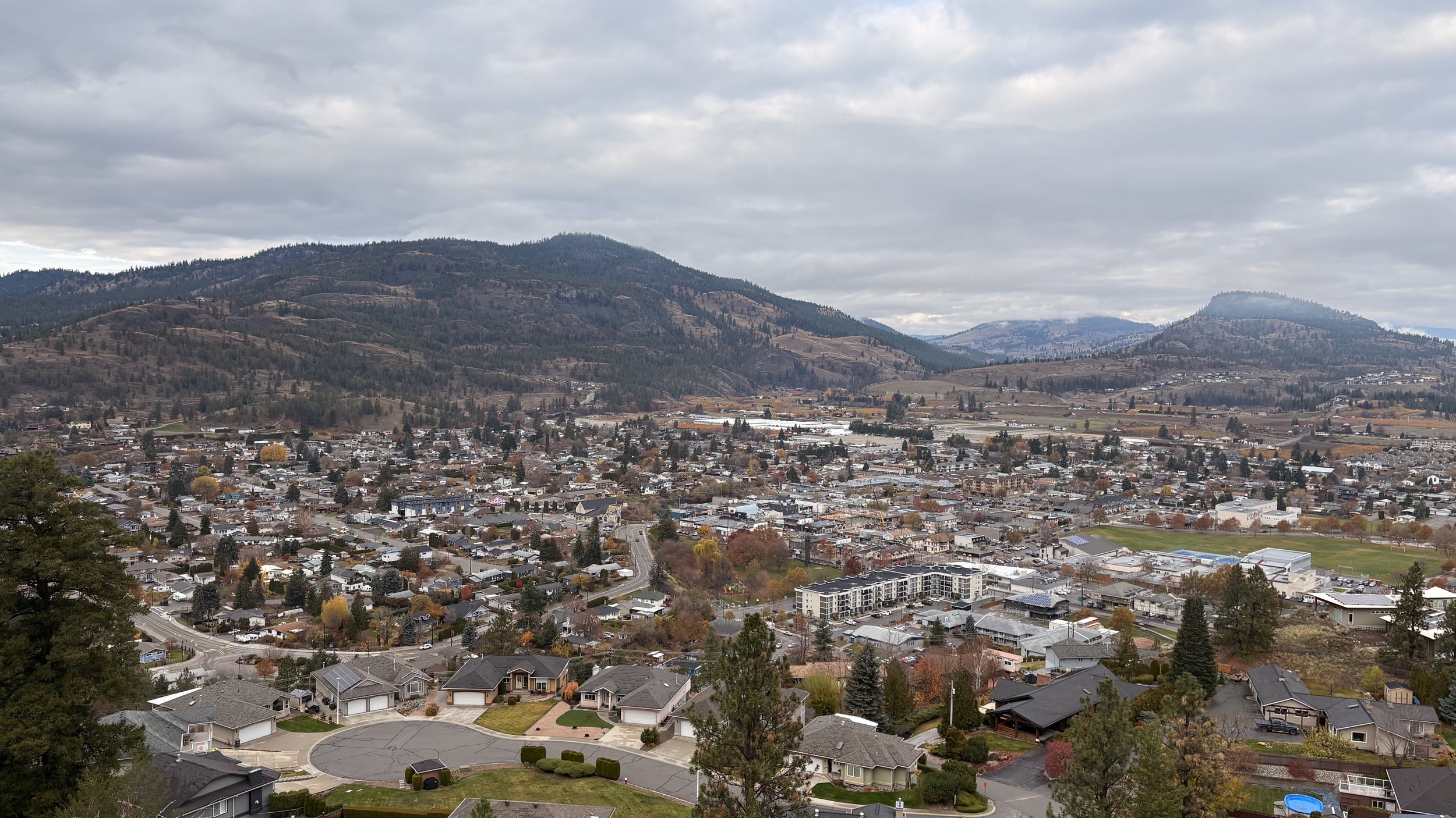 A small city or town with houses, buildings, and streets at the base of surrounding mountains under a cloudy sky.