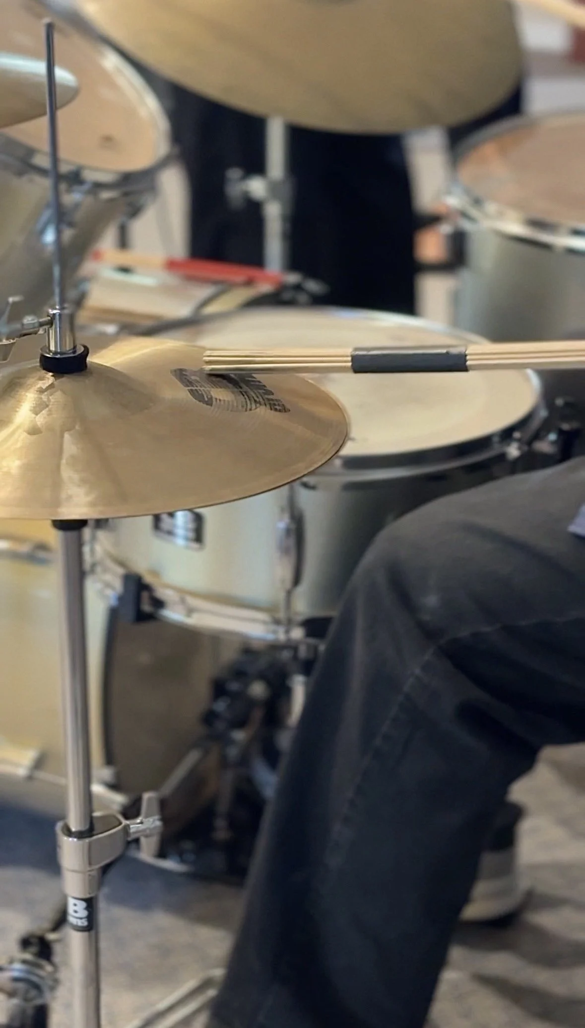 Close-up of a drum set, including cymbals and drum shells, with a person wearing black pants in the background.