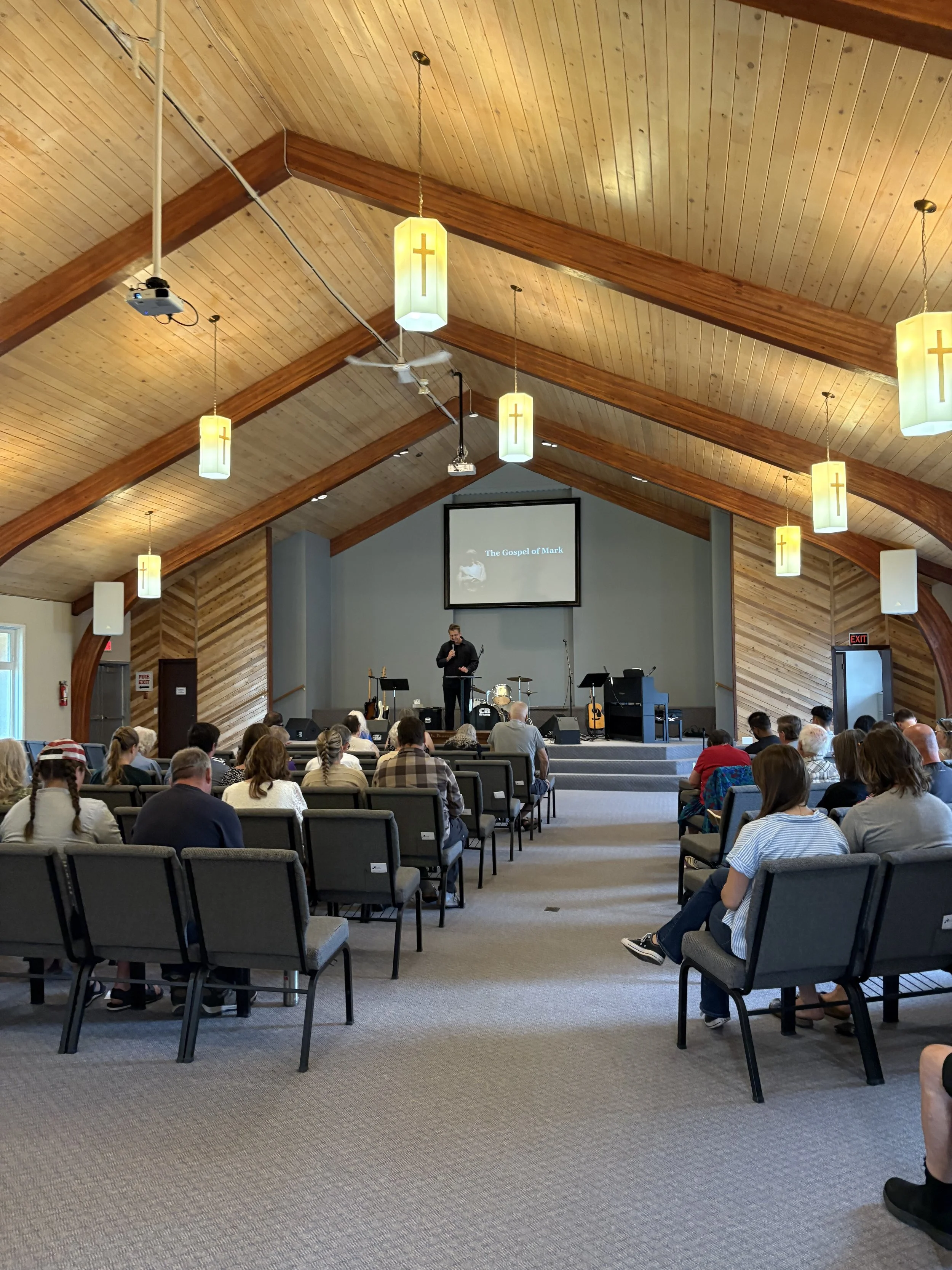 Inside a church with wooden vaulted ceiling, hanging lamps with crosses, and a congregation listening to a speaker on stage. A large screen displays 'The Gospel of Mark'.