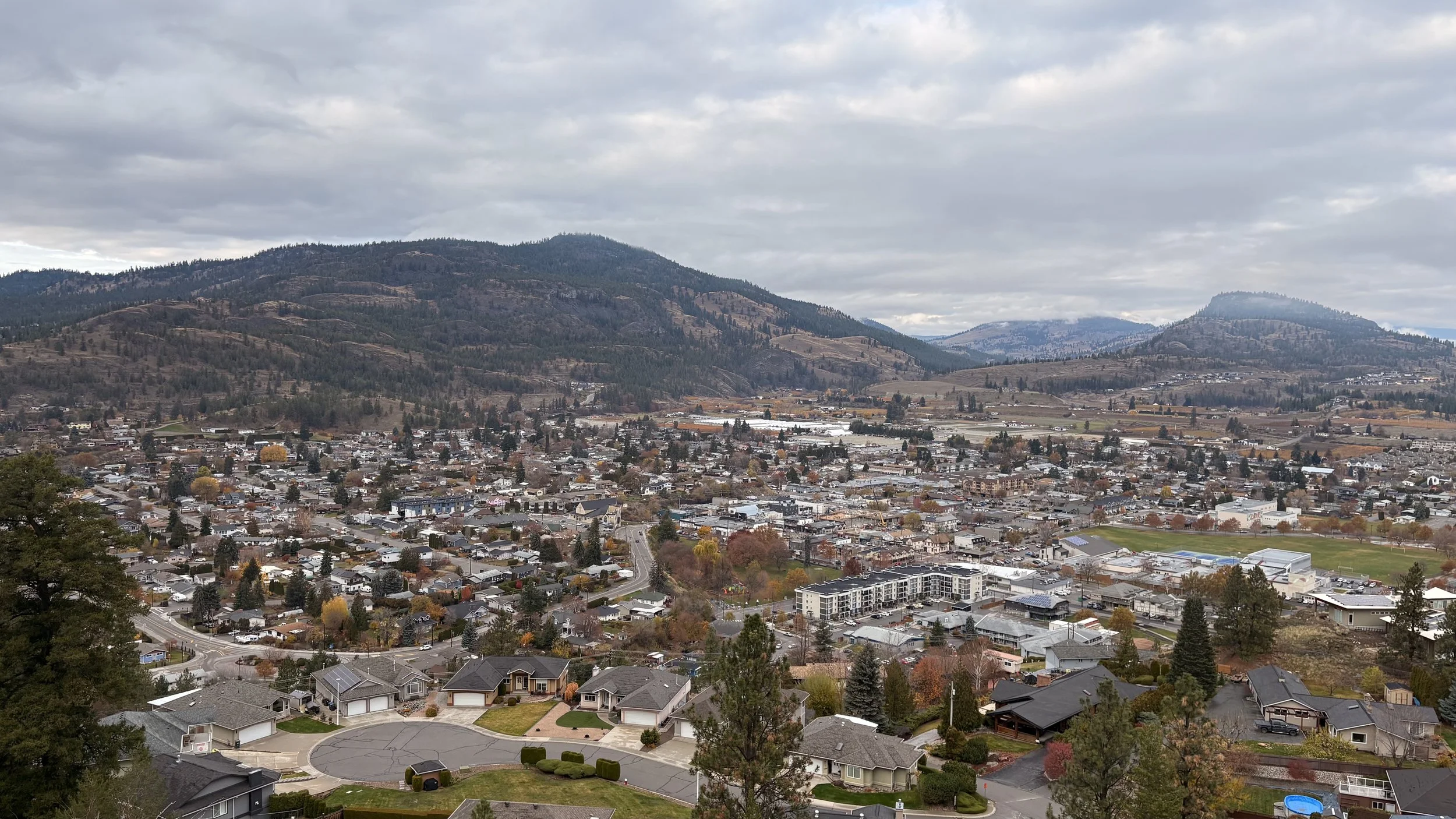 A panoramic view of a small town nestled in a valley surrounded by rolling hills and mountains under a cloudy sky.