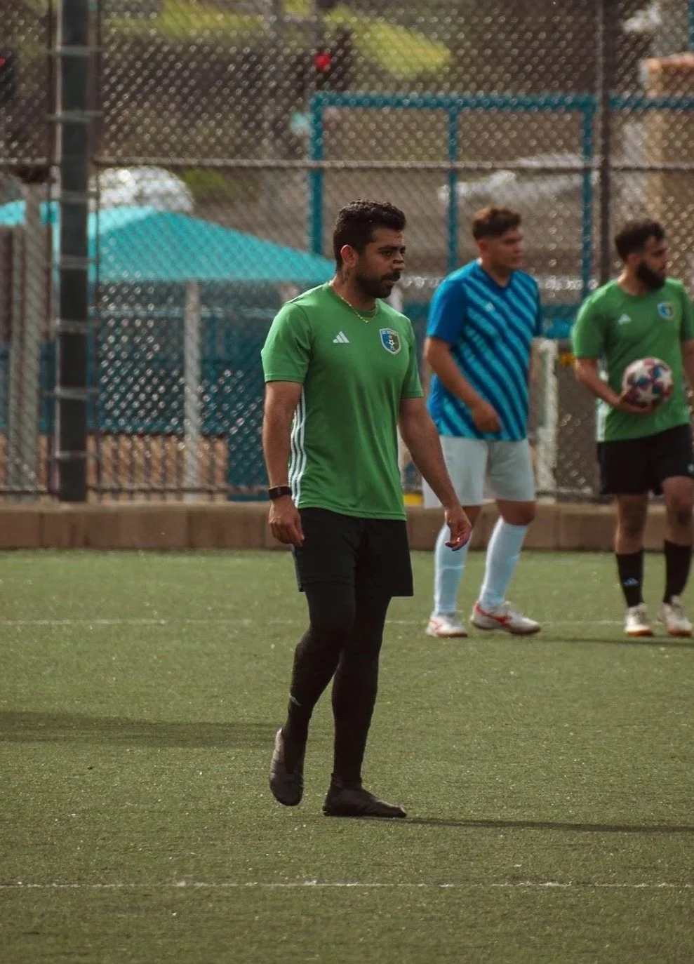 A man in a green soccer jersey and black pants walking on a soccer field, with two other men in sports gear, one holding a soccer ball, in the background.