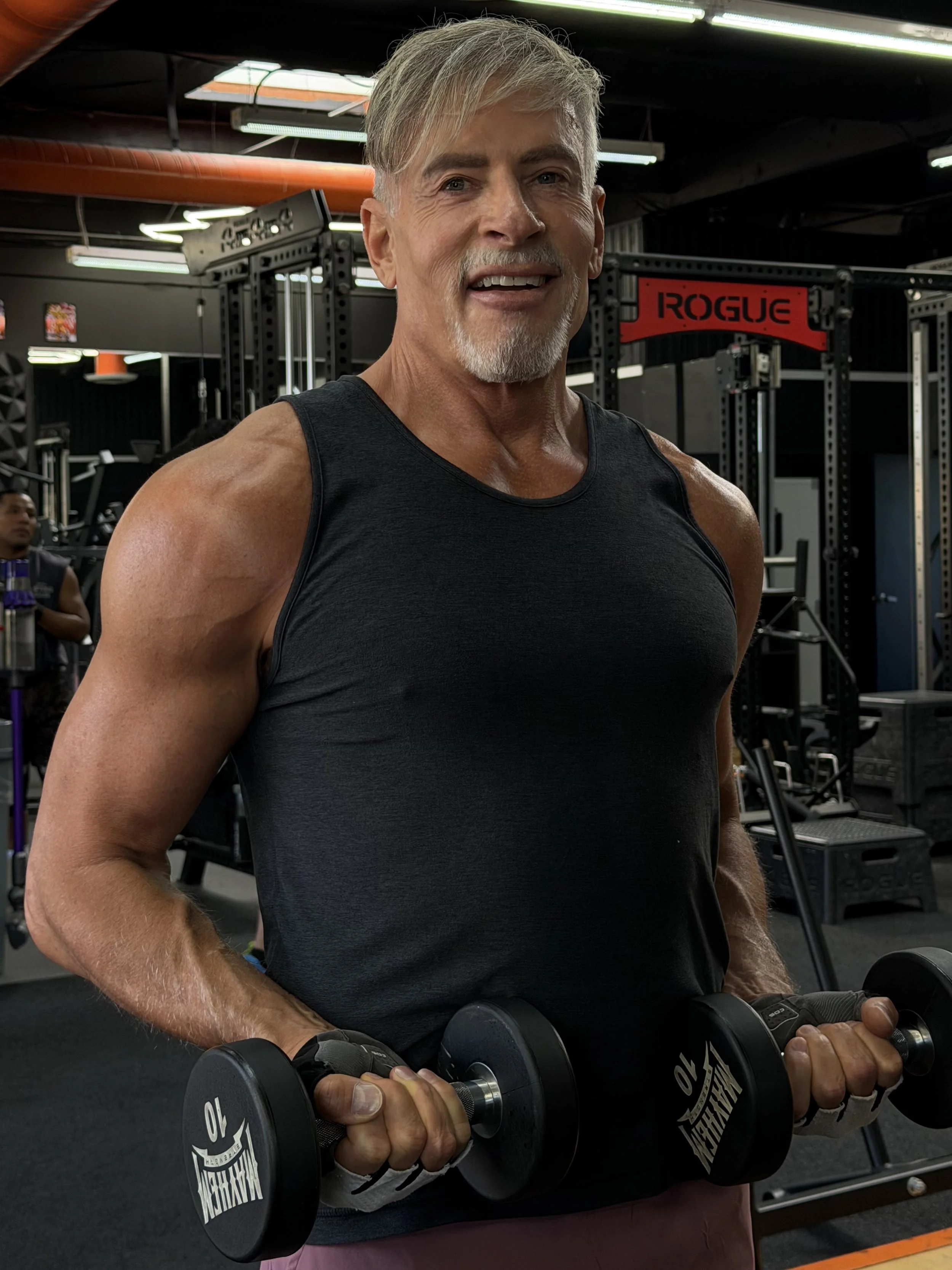 An older man with gray hair and beard lifting dumbbells in a gym.