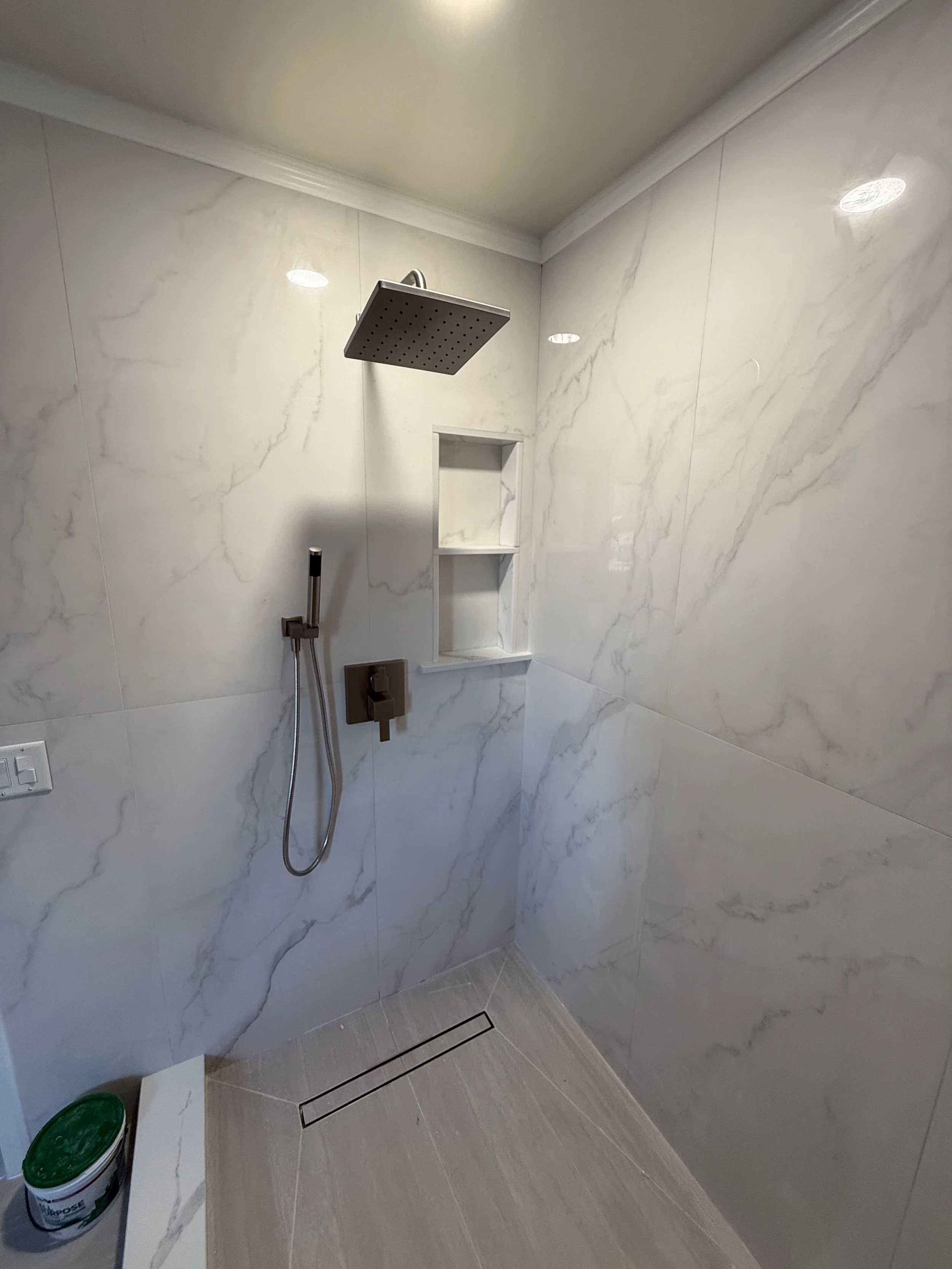 Luxury walk-in shower featuring a rainfall shower head, handheld shower, marble-look porcelain tile, and custom bathroom renovation by Edilgjoni LLC in Rochester, Michigan.