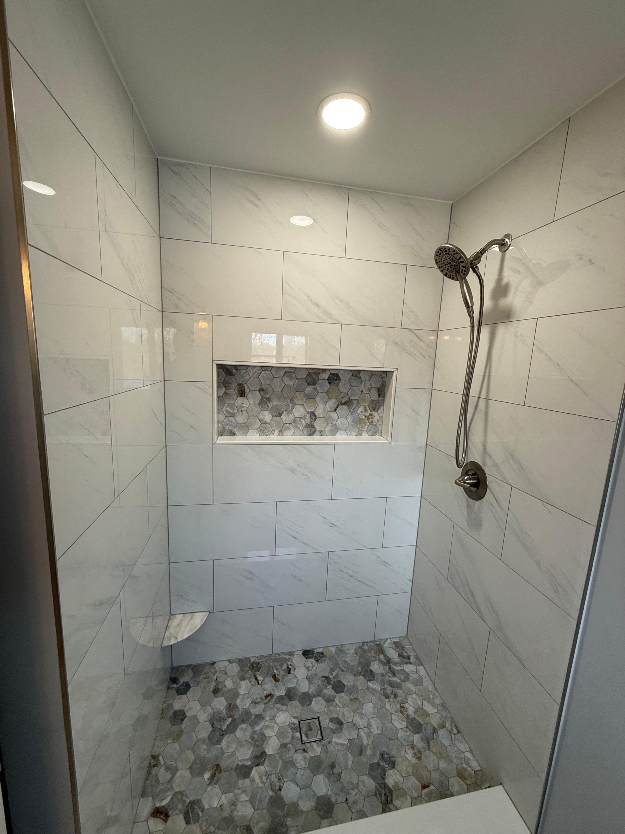 Shower with large white marble tiles on the walls, a built-in niche with hexagonal mosaic tiles, and a hexagonal stone tile floor. There is a silver showerhead and a ceiling light.
