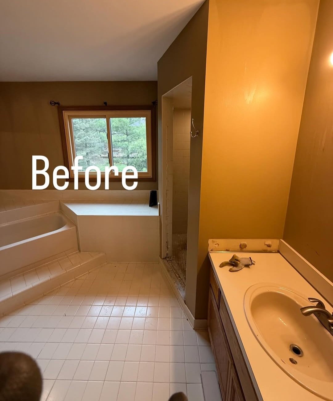 A bathroom with a corner bathtub, a window showing trees outside, an unfinished shower area, a yellow wall, and a sink with gloves on the countertop, with the word 'Before' overlaid.