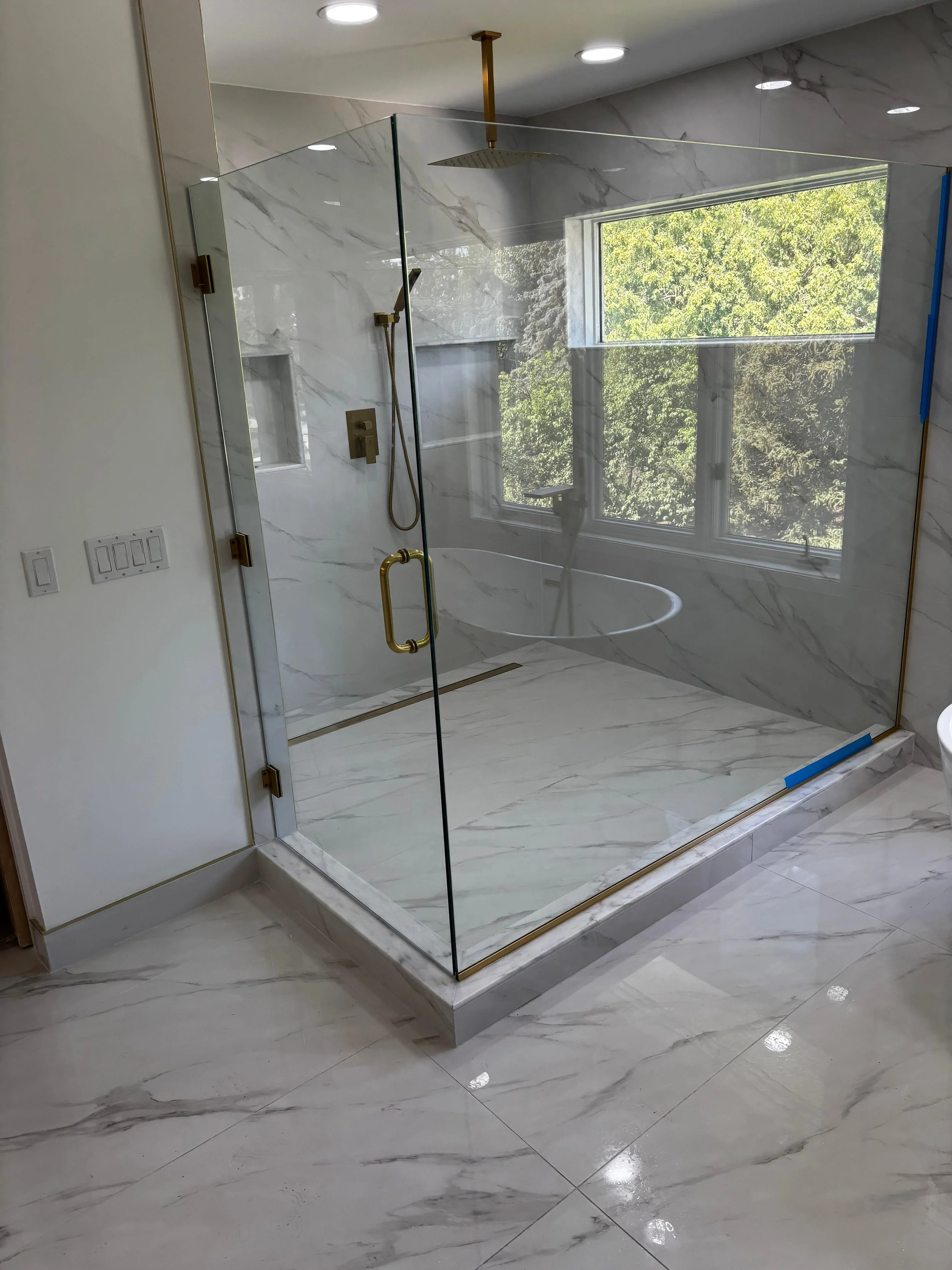 Modern marble bathroom with a glass shower enclosure, large window, and gold fixtures.