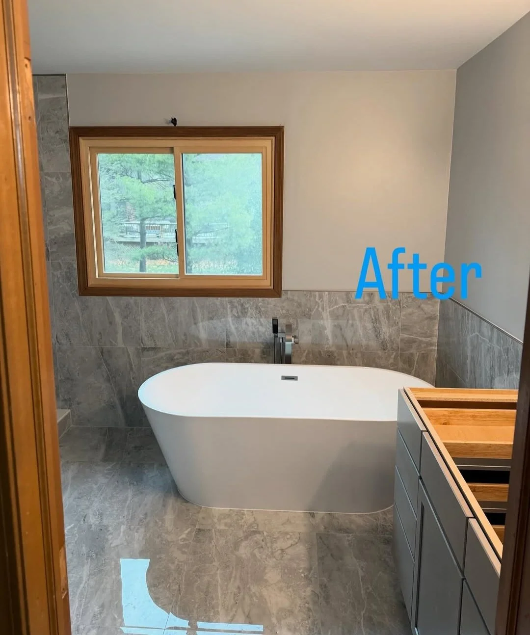 A bathroom with a freestanding bathtub, a window above the tub, and a vanity with a wooden tray. The walls have grey marble tiles and the ceiling is painted white.