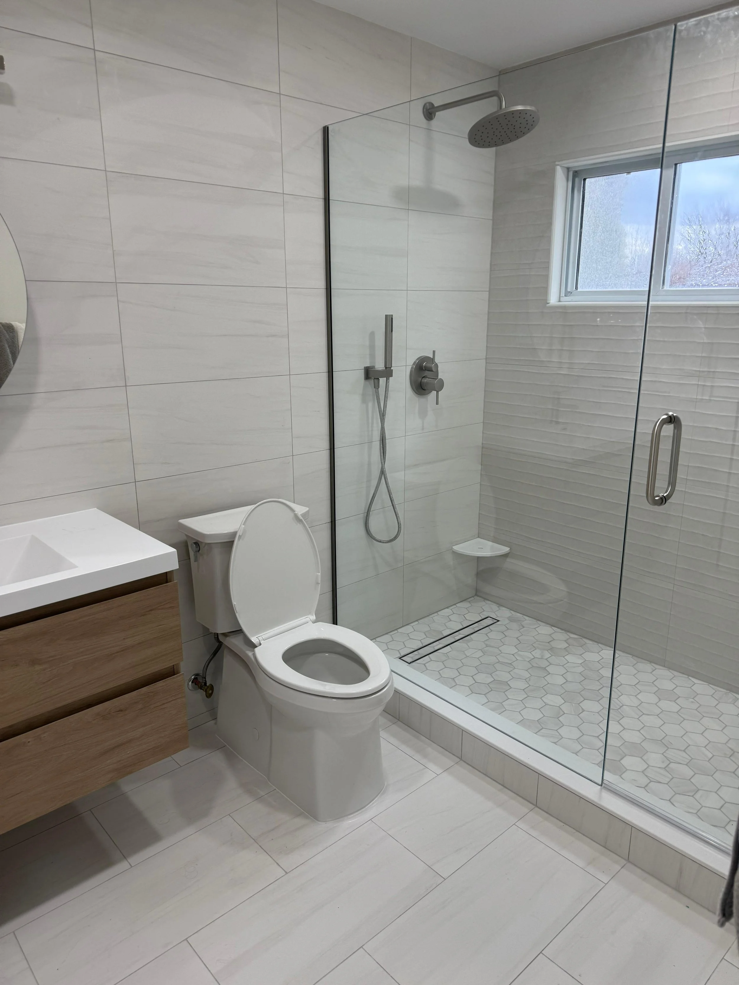 Modern bathroom with a walk-in shower, toilet, and vanity with a sink. The shower has a rainfall showerhead and a window. The floor and walls are tiled with light-colored tiles.