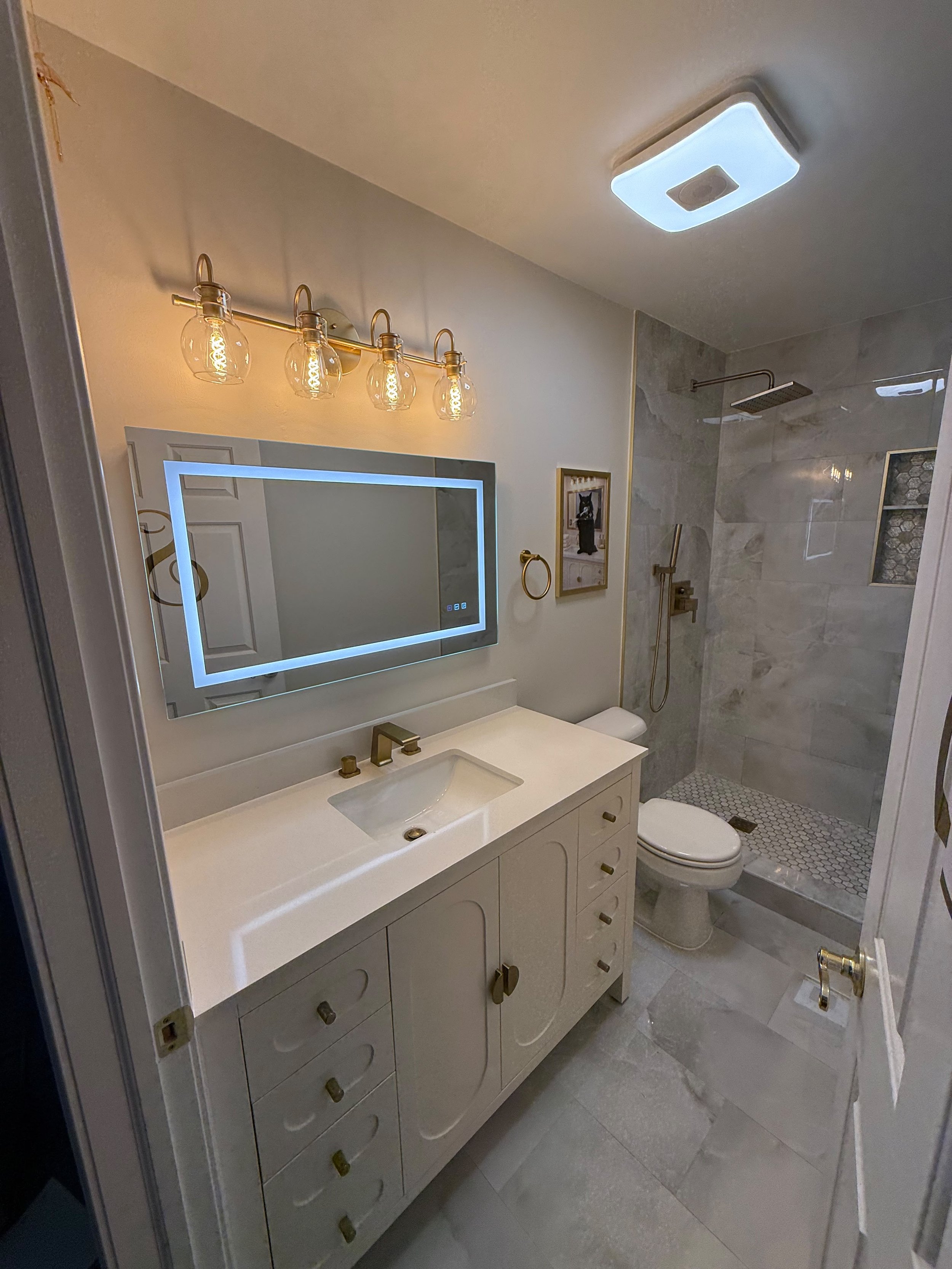 Custom bathroom vanity with a quartz countertop, LED mirror, warm vanity lighting, and an open view of the tiled walk-in shower.