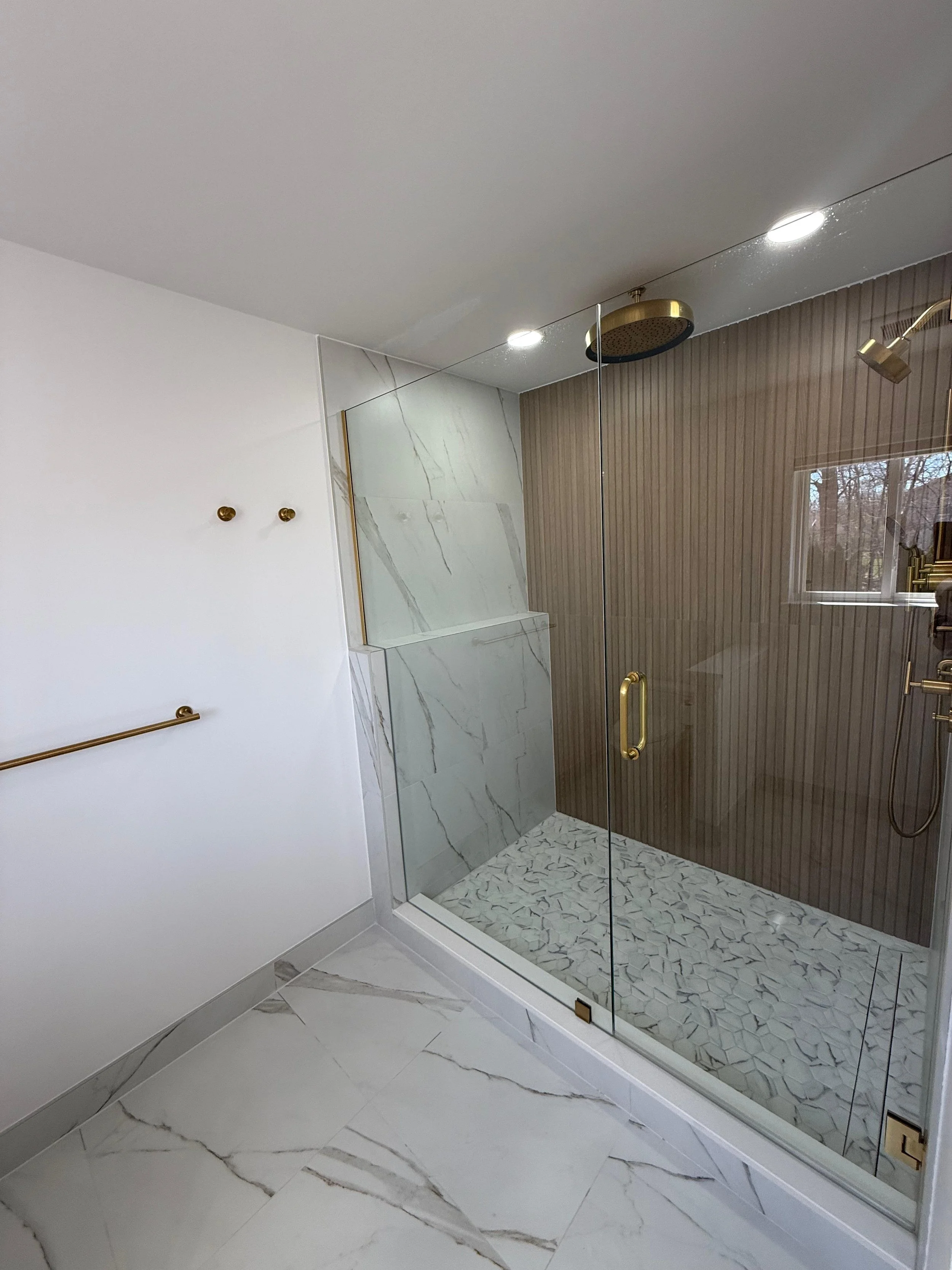 Modern bathroom shower area with marble and wood textures, glass enclosure, brass fixtures, and white marble flooring.
