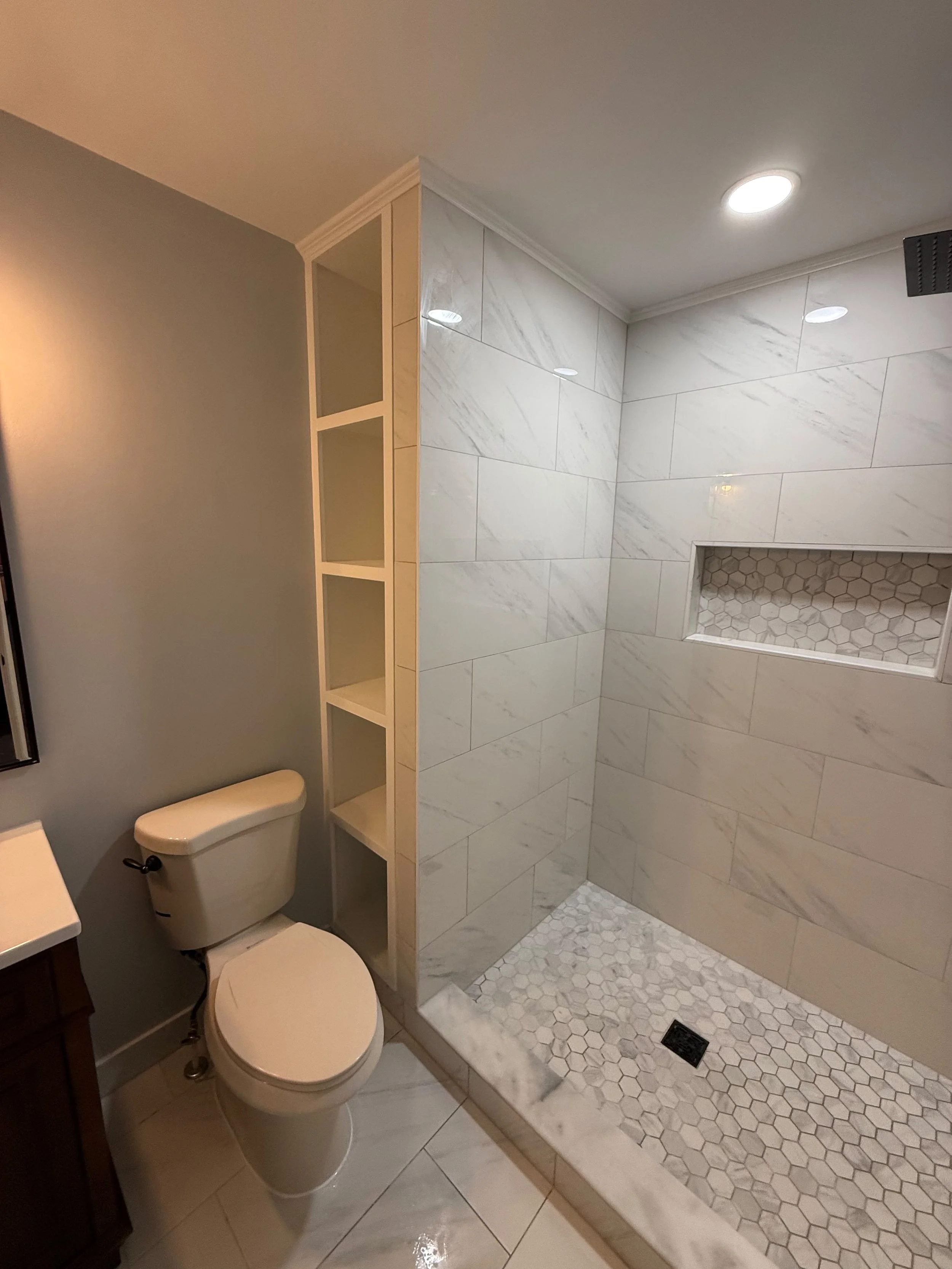 Complete bathroom remodel showcasing a curbless walk-in shower, floating storage shelves, modern toilet, gray porcelain tile, and a spacious contemporary layout in Rochester , MI.