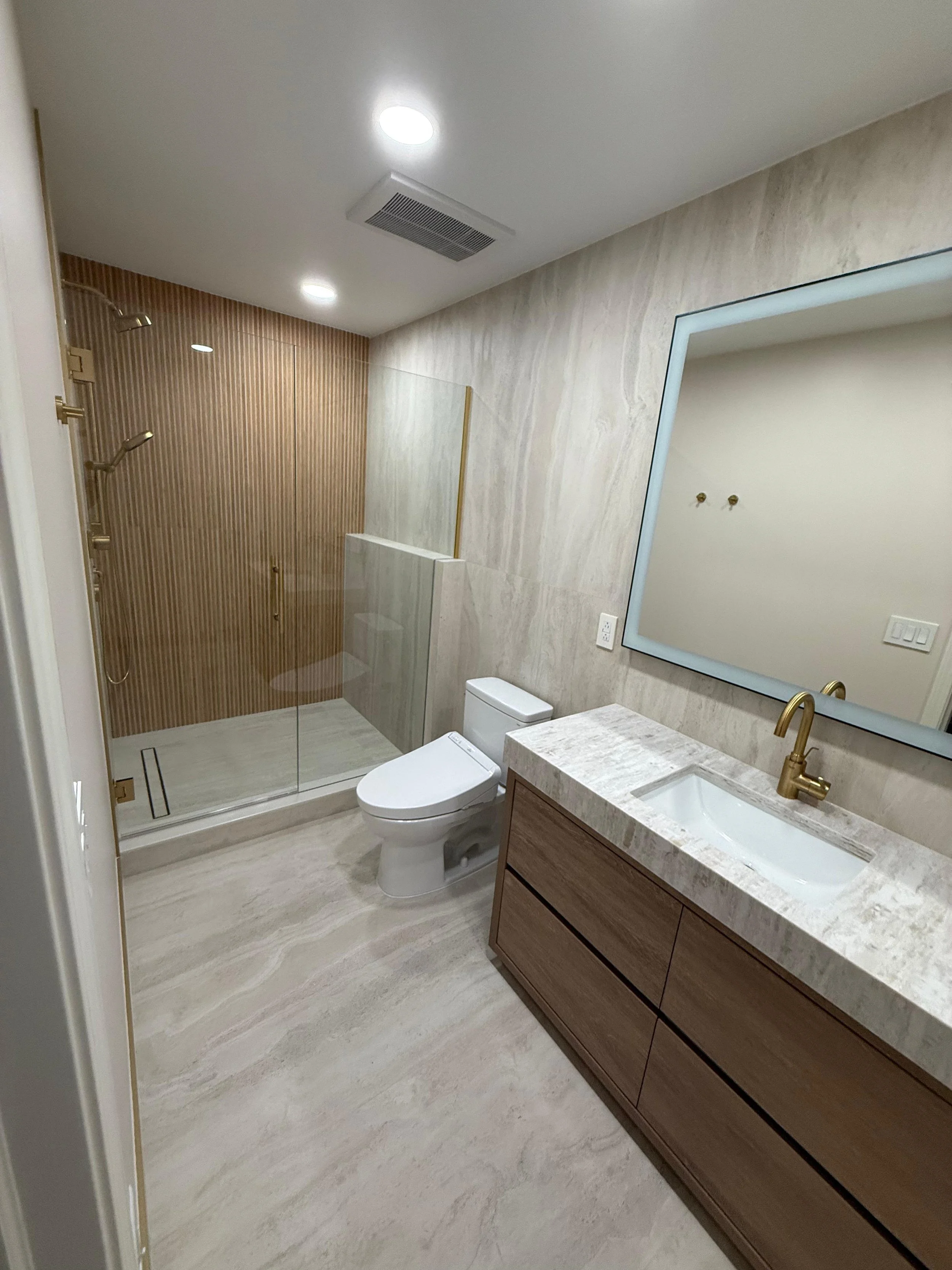 Modern bathroom with a shower with a glass enclosure, a toilet, a vanity with a marble countertop and a mirror, and beige marble flooring and walls.