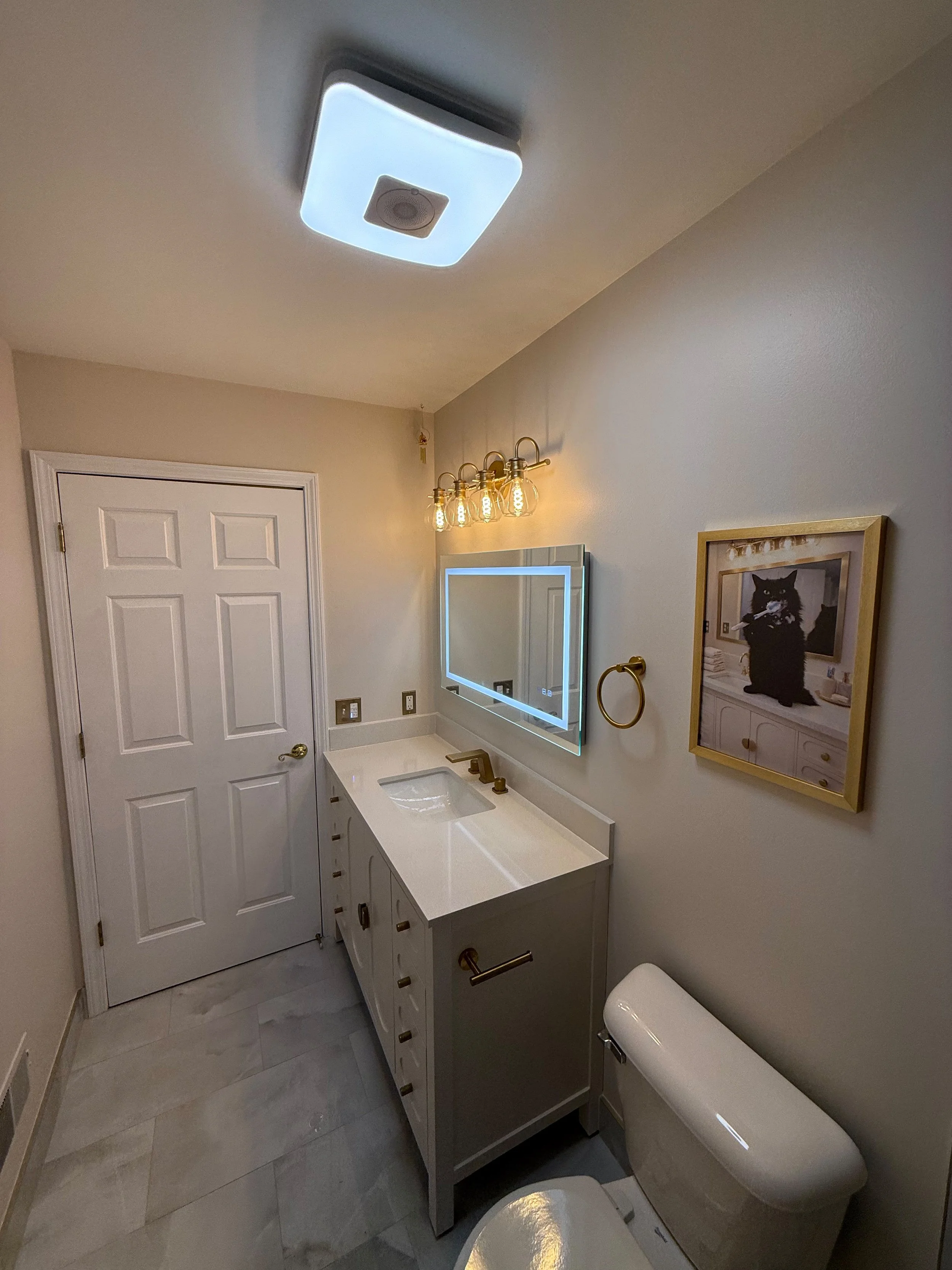 Modern bathroom with a white single-sink vanity, illuminated mirror, brushed nickel faucet, porcelain floor tile, and warm vanity lighting in Rochester, MI.
