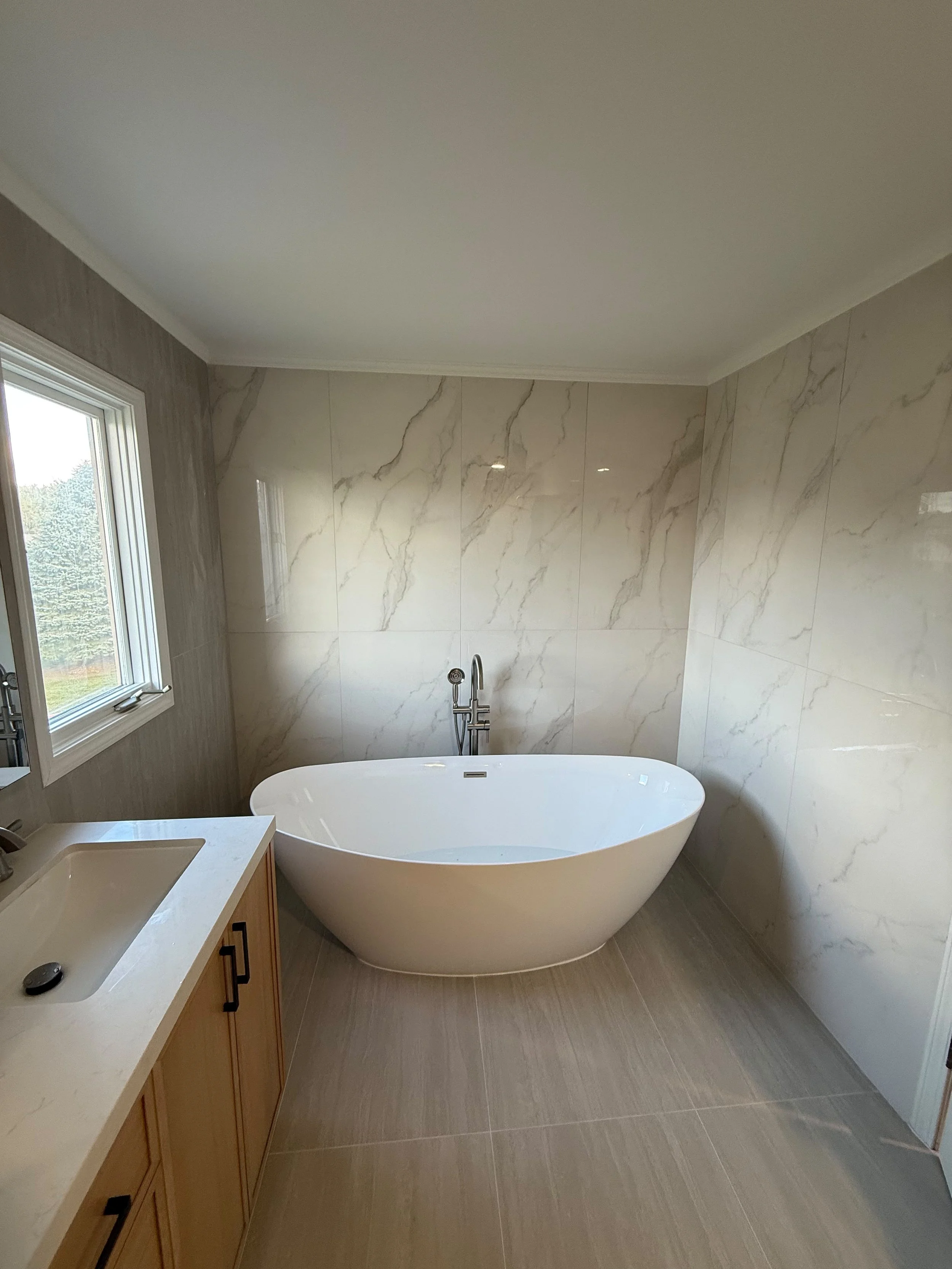 Luxury bathroom remodel featuring a freestanding soaking tub, large marble-look wall tile, modern wood vanity, and premium tile flooring completed by Edilgjoni LLC in Rochester, Michigan.