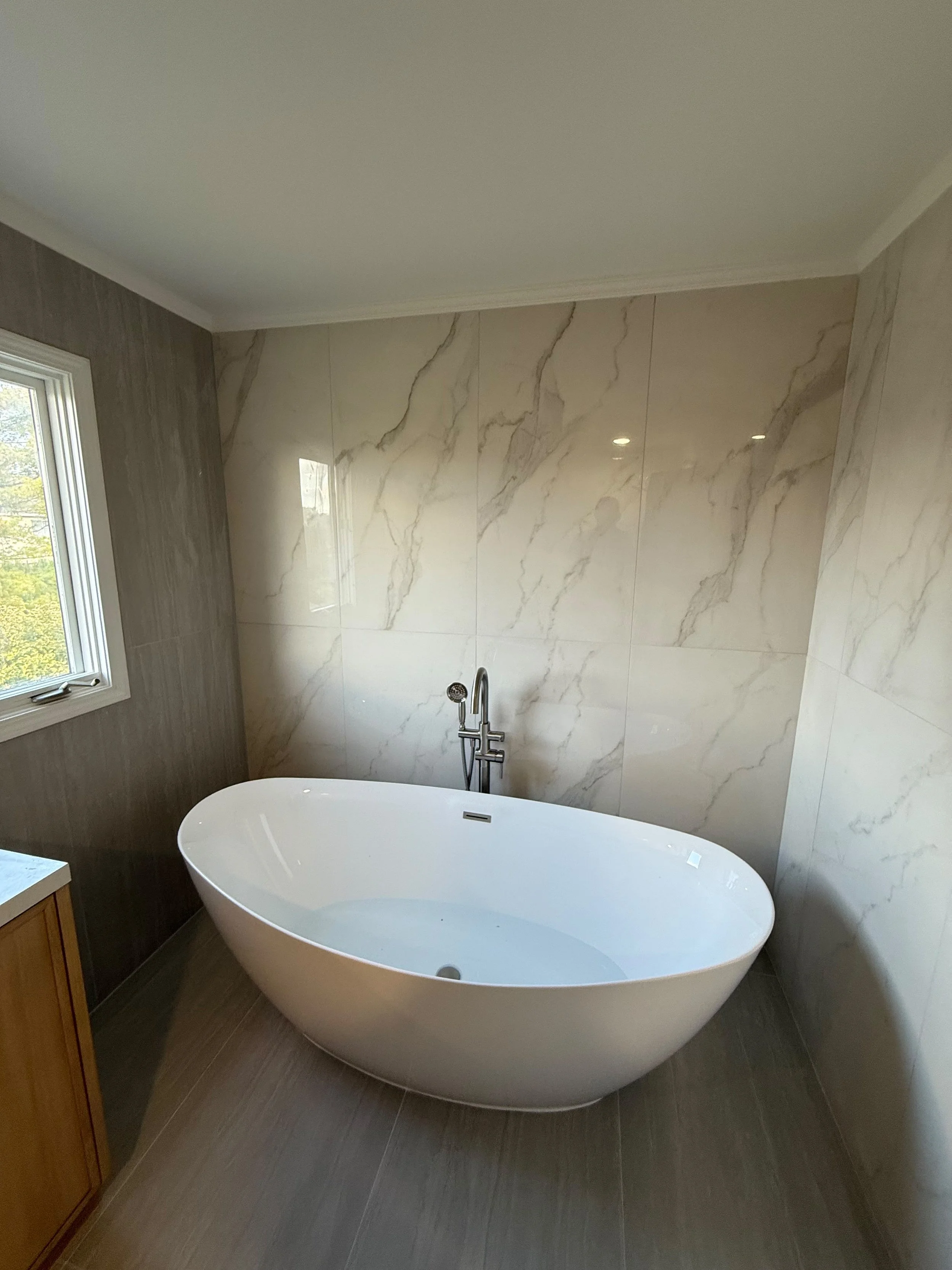 Close-up view of a freestanding bathtub with floor-mounted tub faucet, marble-look porcelain tile walls, natural light, and modern bathroom design by Edilgjoni LLC in Rochester, Michigan.