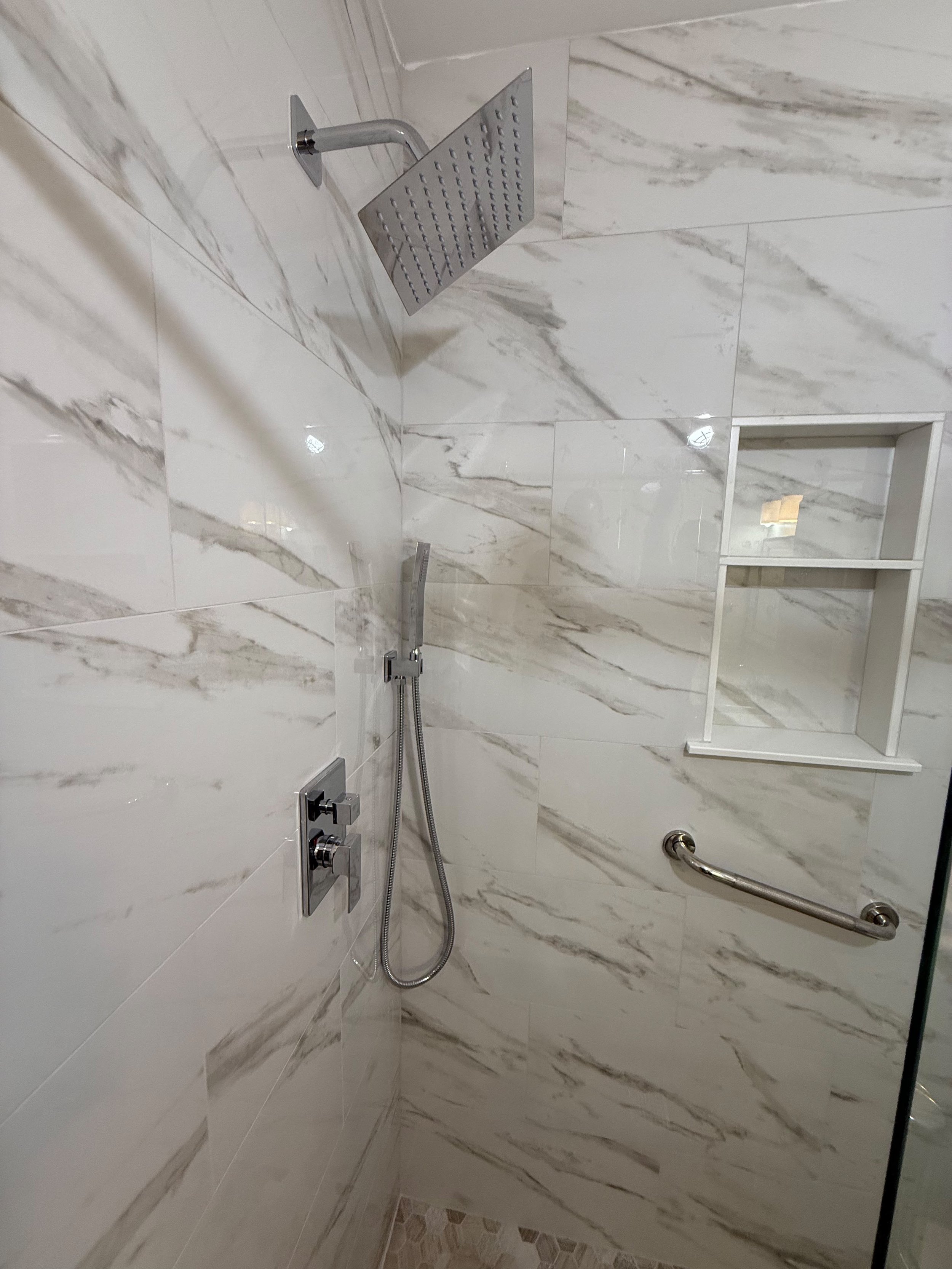 A complete bathroom remodel featuring a spacious walò-in shower, marble-look porcelain tile, custom glass enclosure, recessed shelving, and high-quality craftsmanship that creates a clean, luxurious space.