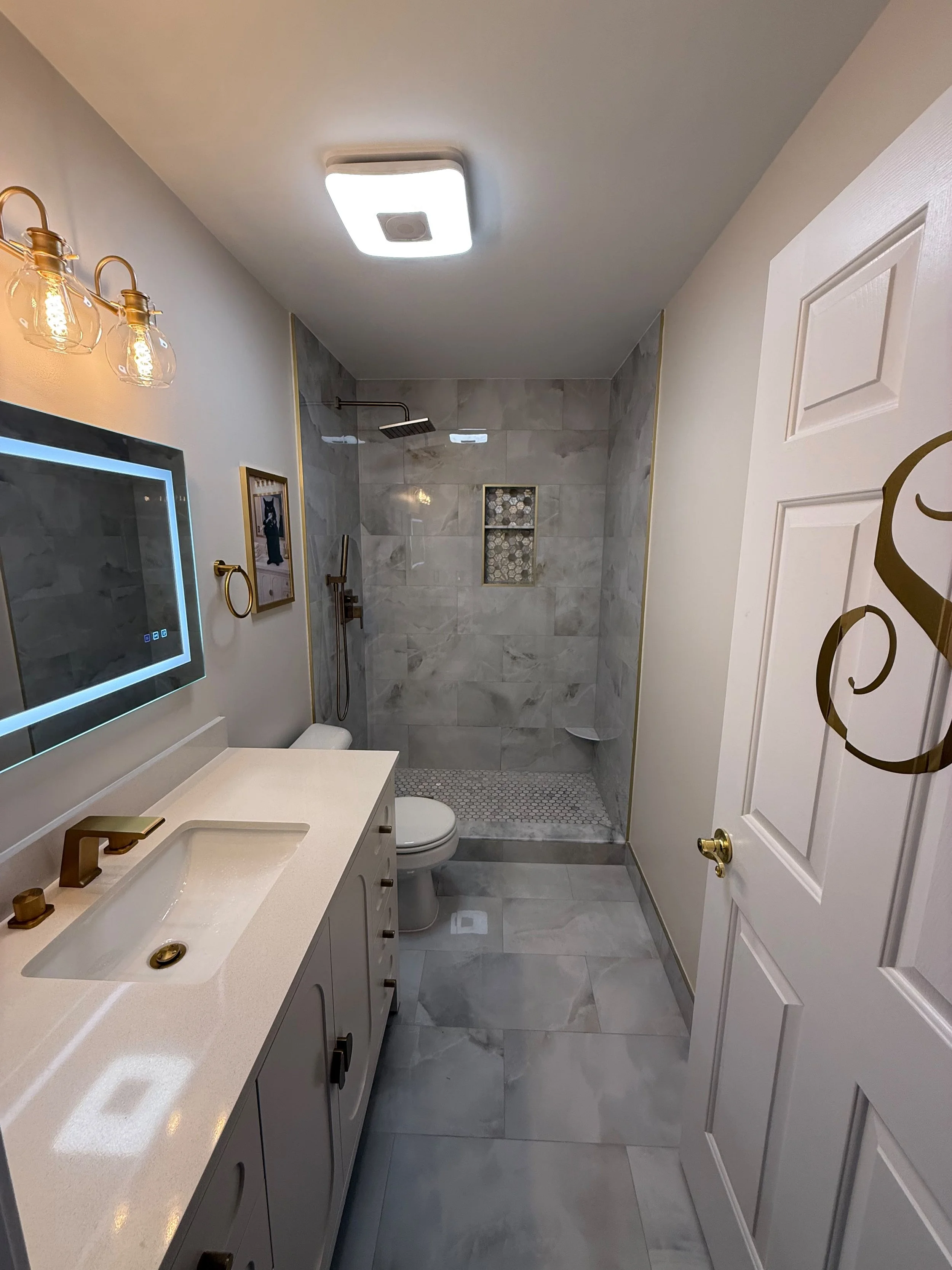 Modern bathroom with white vanity, gold fixtures, marble walls and floor, shower with niche and rainfall showerhead, backlit mirror, and decorative wall art.