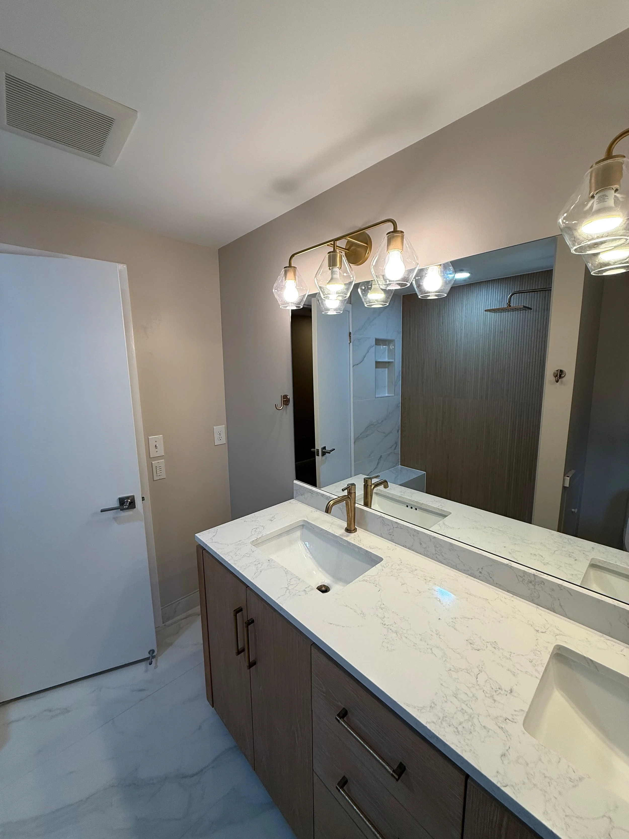 Custom double - sink vanity with quartz  countertop, brushed nickel faucets, oversized framed mirror, warm wood cabinetry, and decorative vanity lighting in a newly remodel bathroom.