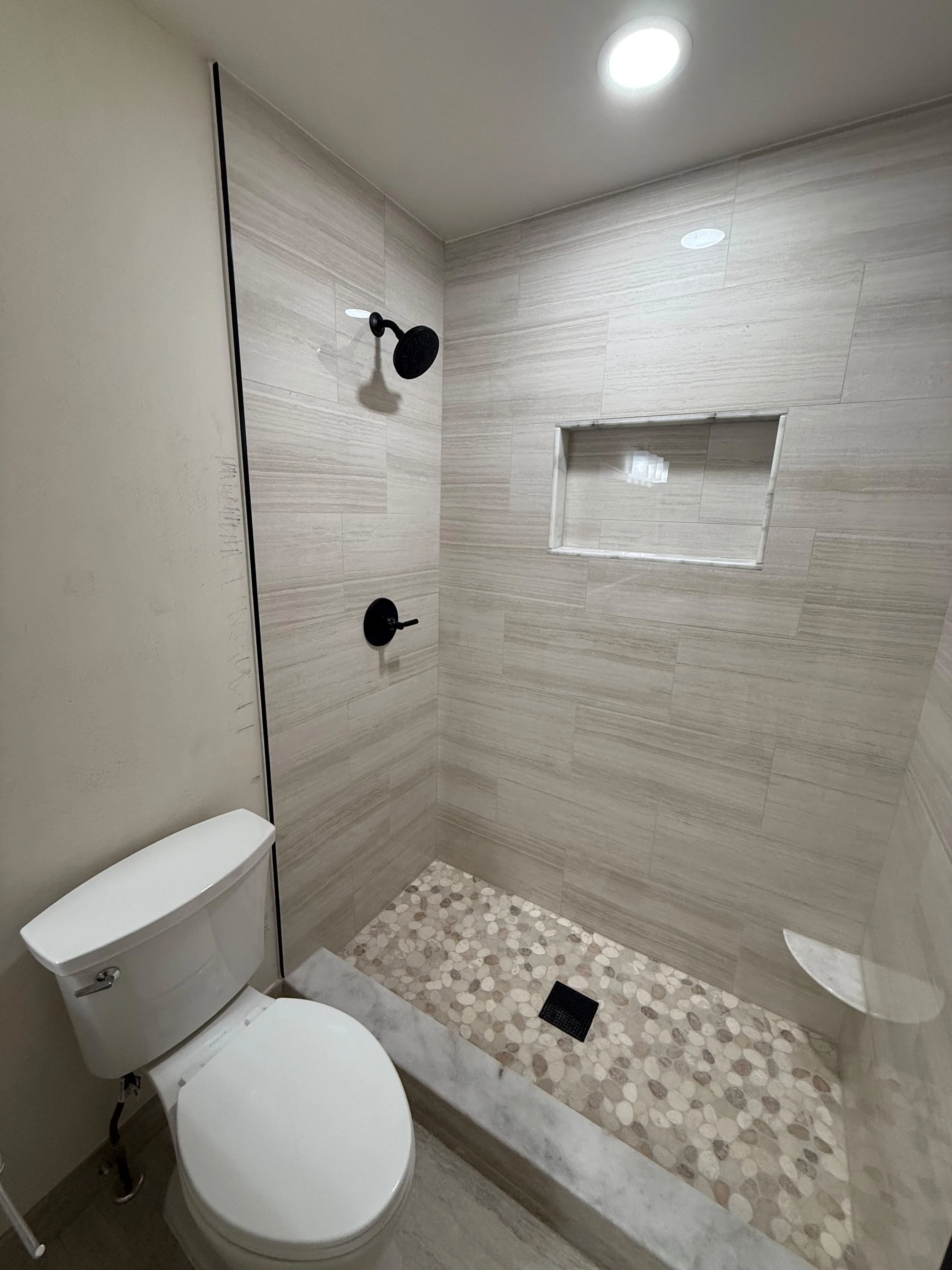 Bathroom remodel with custom walk-in shower, white tile walls. pebble shower floor, built-in niche, and matte black fixtures by Edilgjoni LLC in Southeast Michigan.