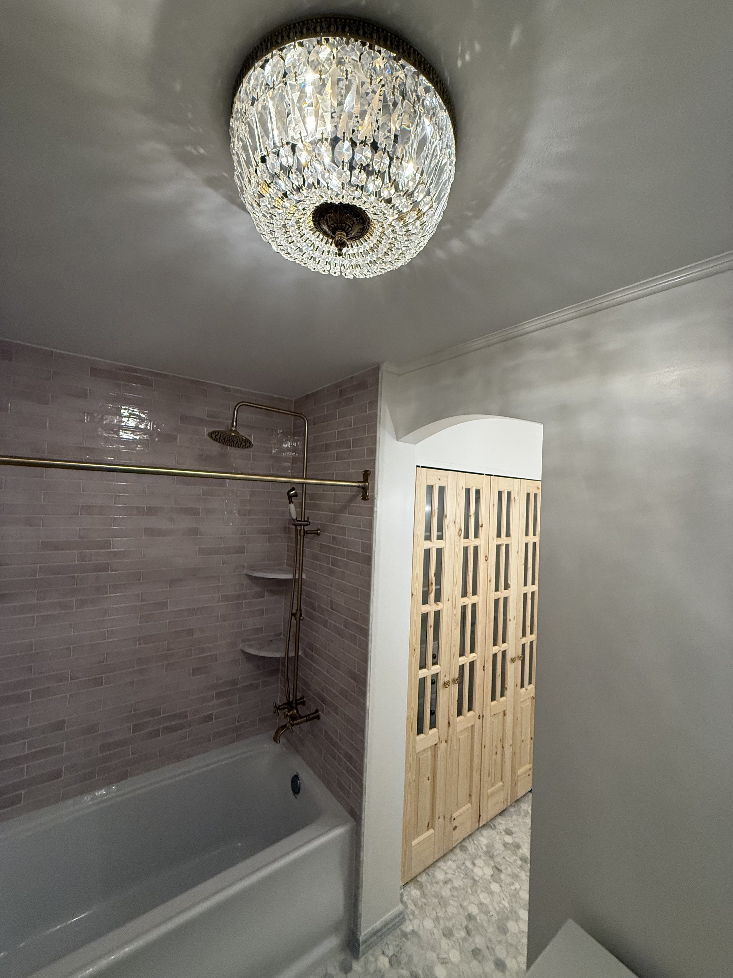 Completed bathroom featuring a decorative ceiling light, gray tiled bathtub surround, and natural wood glass-panel door.