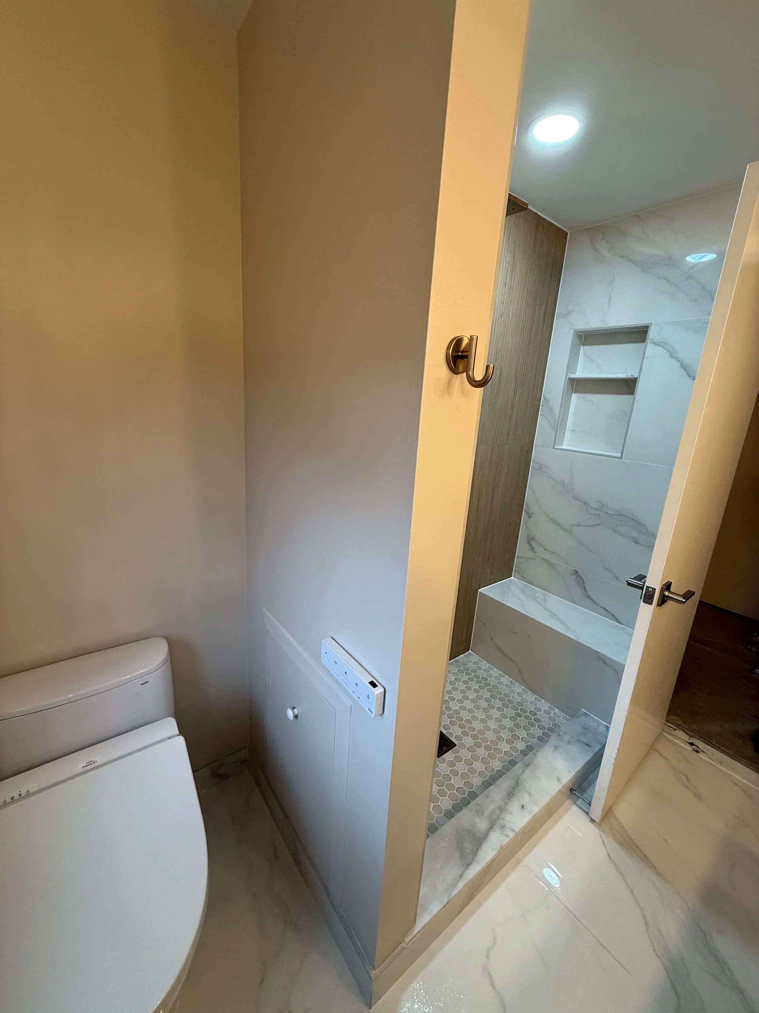 Bright bathroom featuring a private toilet area with a custom tiled walk-in shower, recessed wall niche, pebble shower floor, and soft gray wall finishes in Rochester, MI.