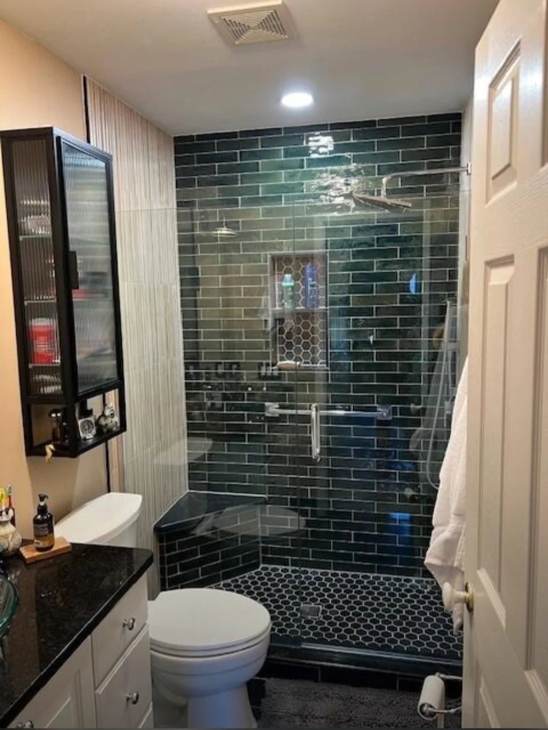 Bathroom with a walk-in shower featuring black subway tiles and hexagonal black and white floor tiles, a white toilet, black countertop with toiletries, and a beige wall with a vertical wood panel, mirror, and cabinet.