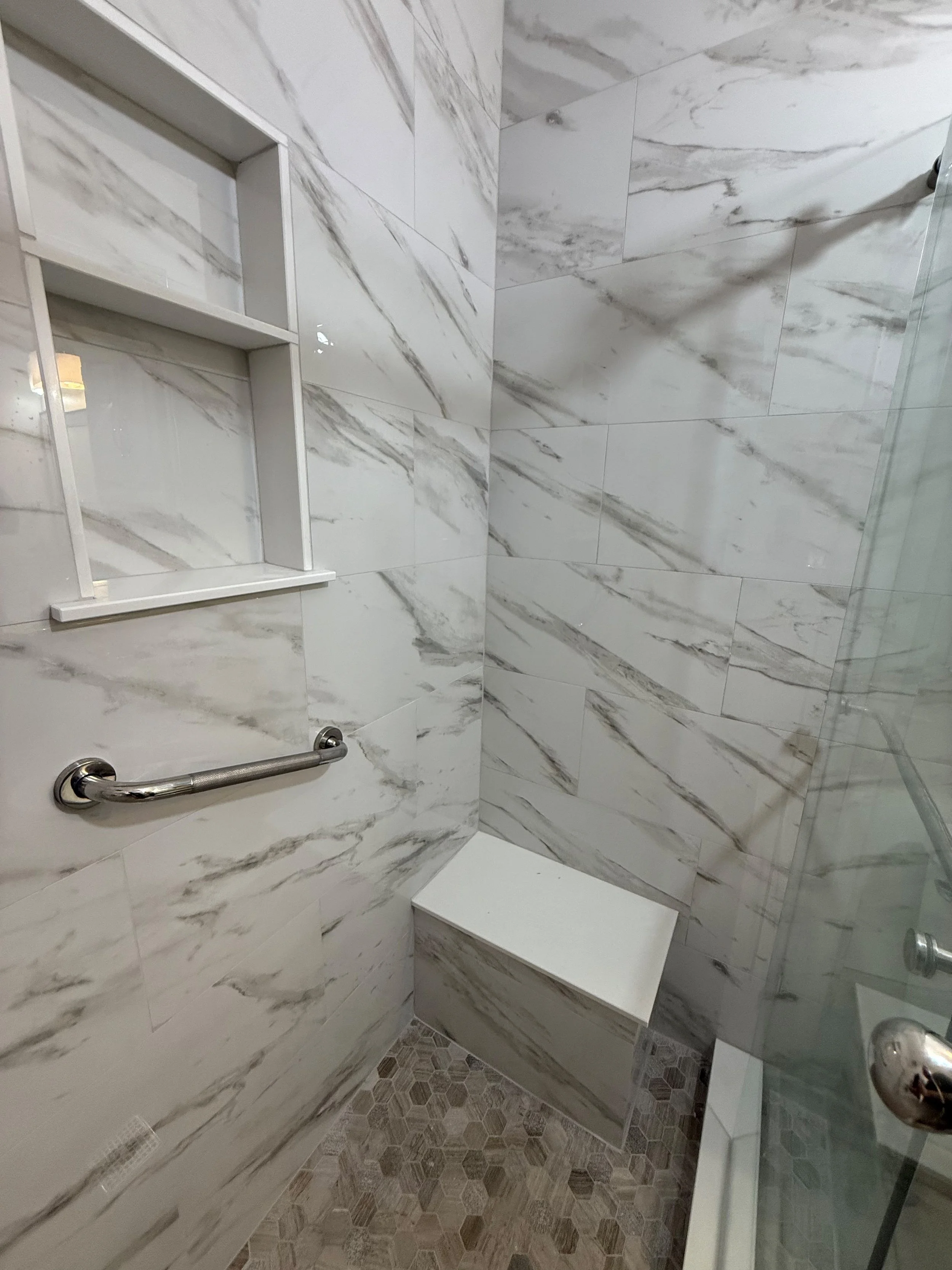 Spa-inspired shower with a floating corner bench, recessed storage, shelves, grab bars, marble-look porcelain tile, and a pebble mosaic shower floor.