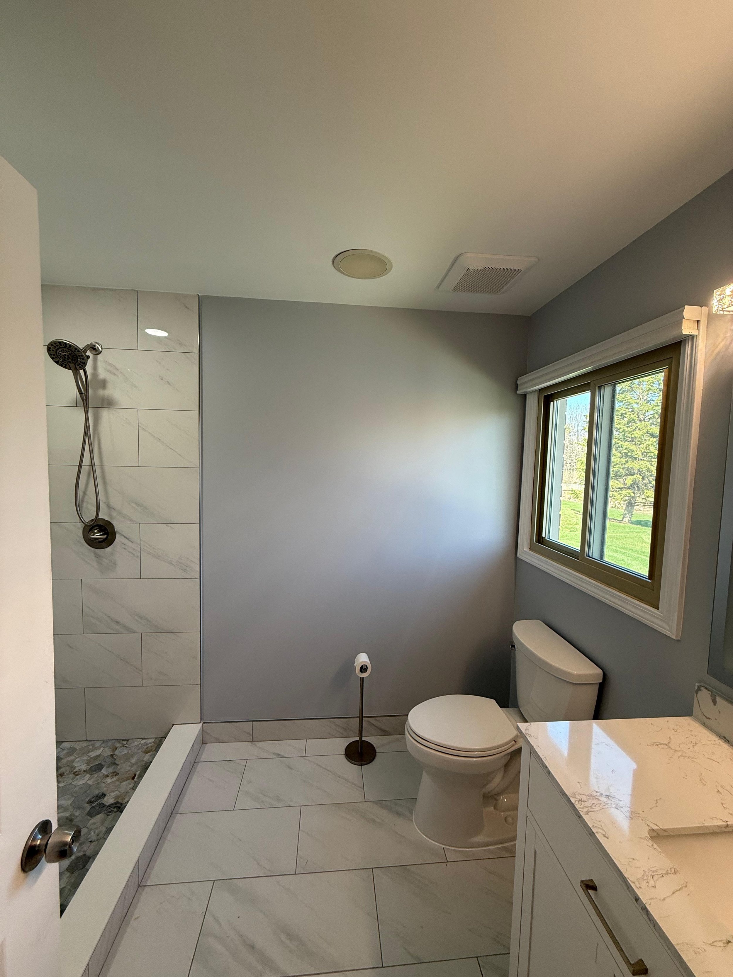 Bathroom with a walk-in shower with marble and tile walls, a showerhead, a toilet, a window showing greenery outside, and a marble countertop.