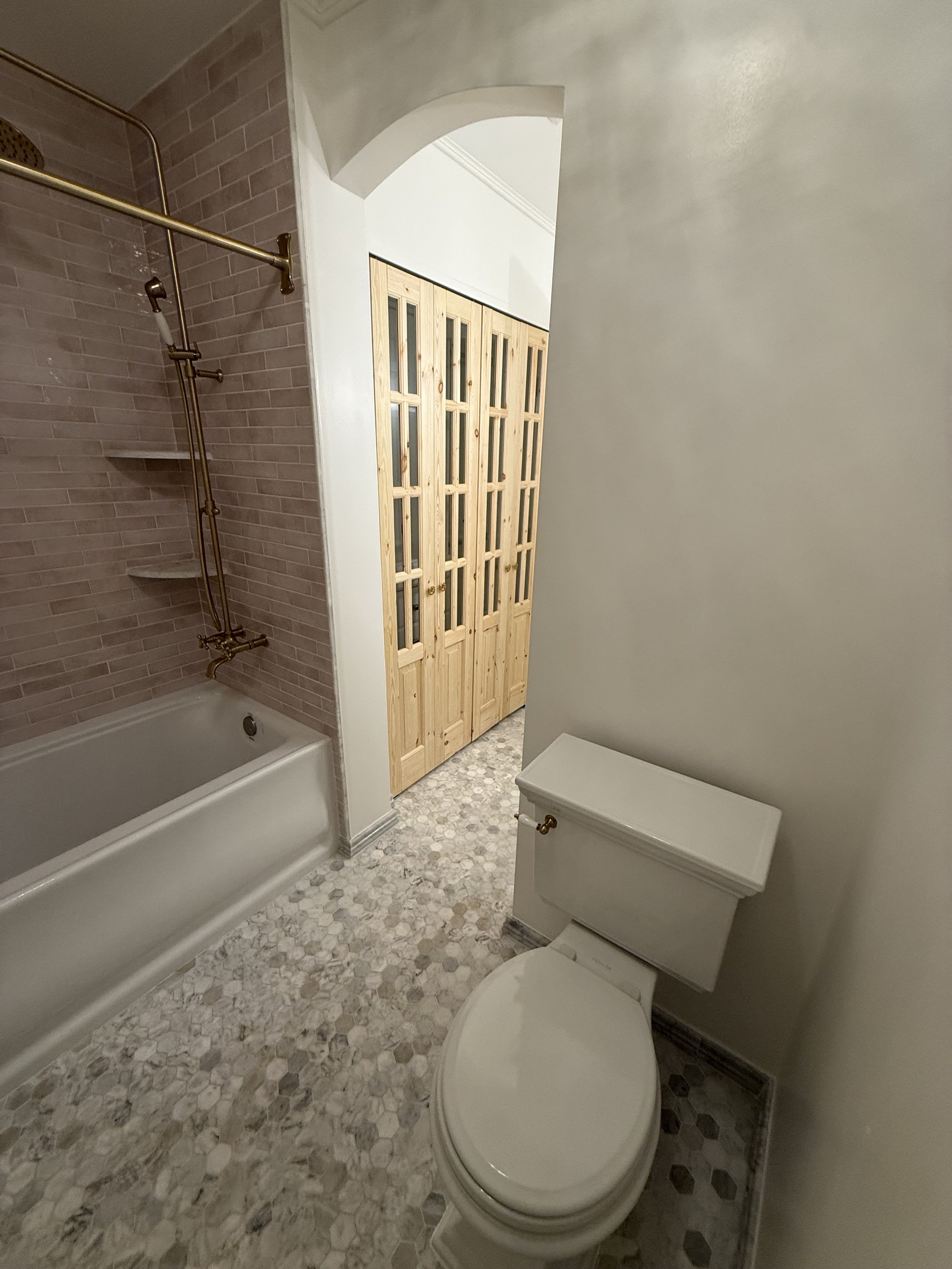 Remodeled bathroom with a white toilet, gray tiled walls, patterned flooring, and a natural wood glass-panel door.