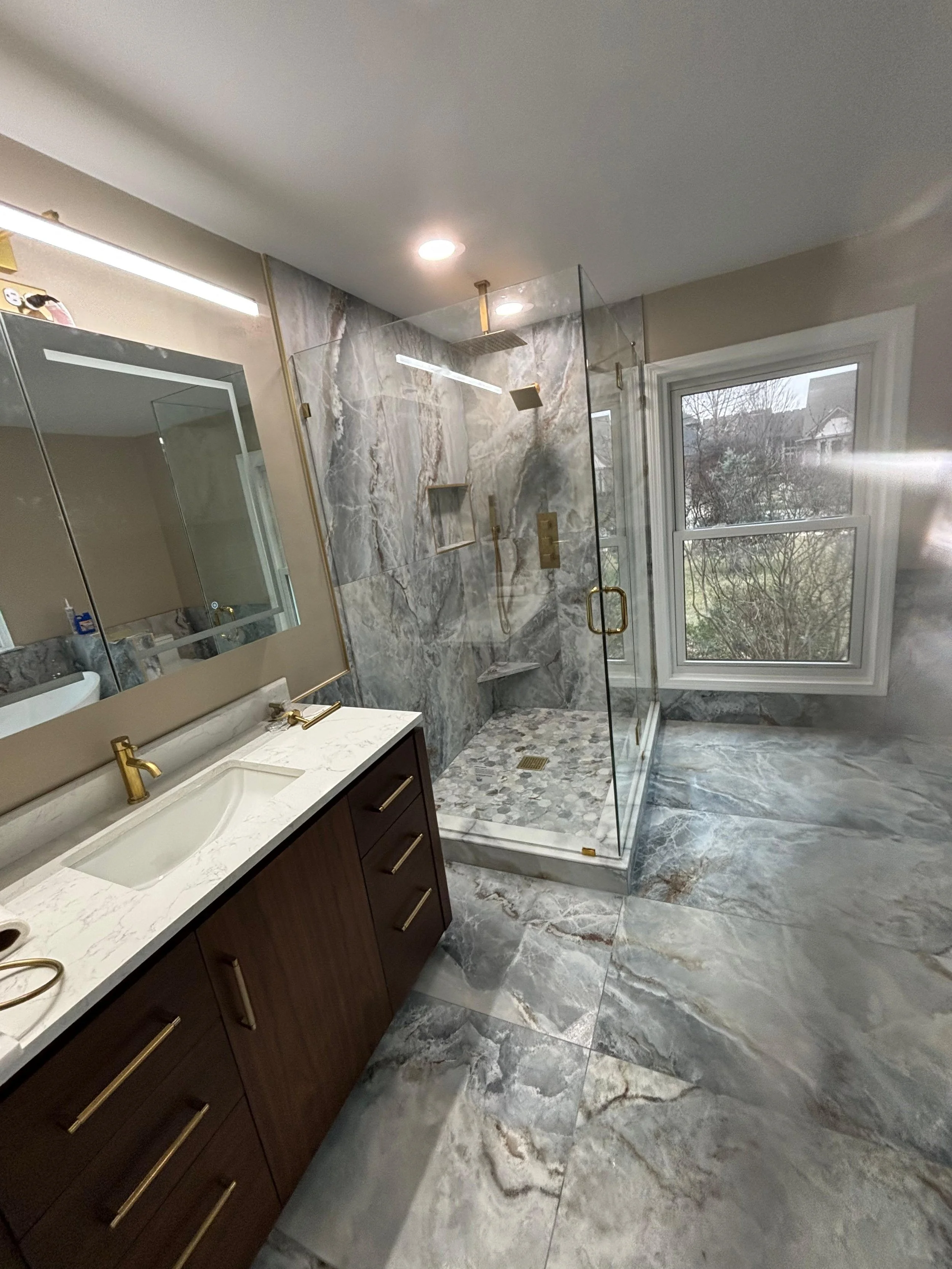 Modern bathroom with a marble shower with glass doors, a window showing an outdoor view, and a vanity with a marble countertop and a mirror.