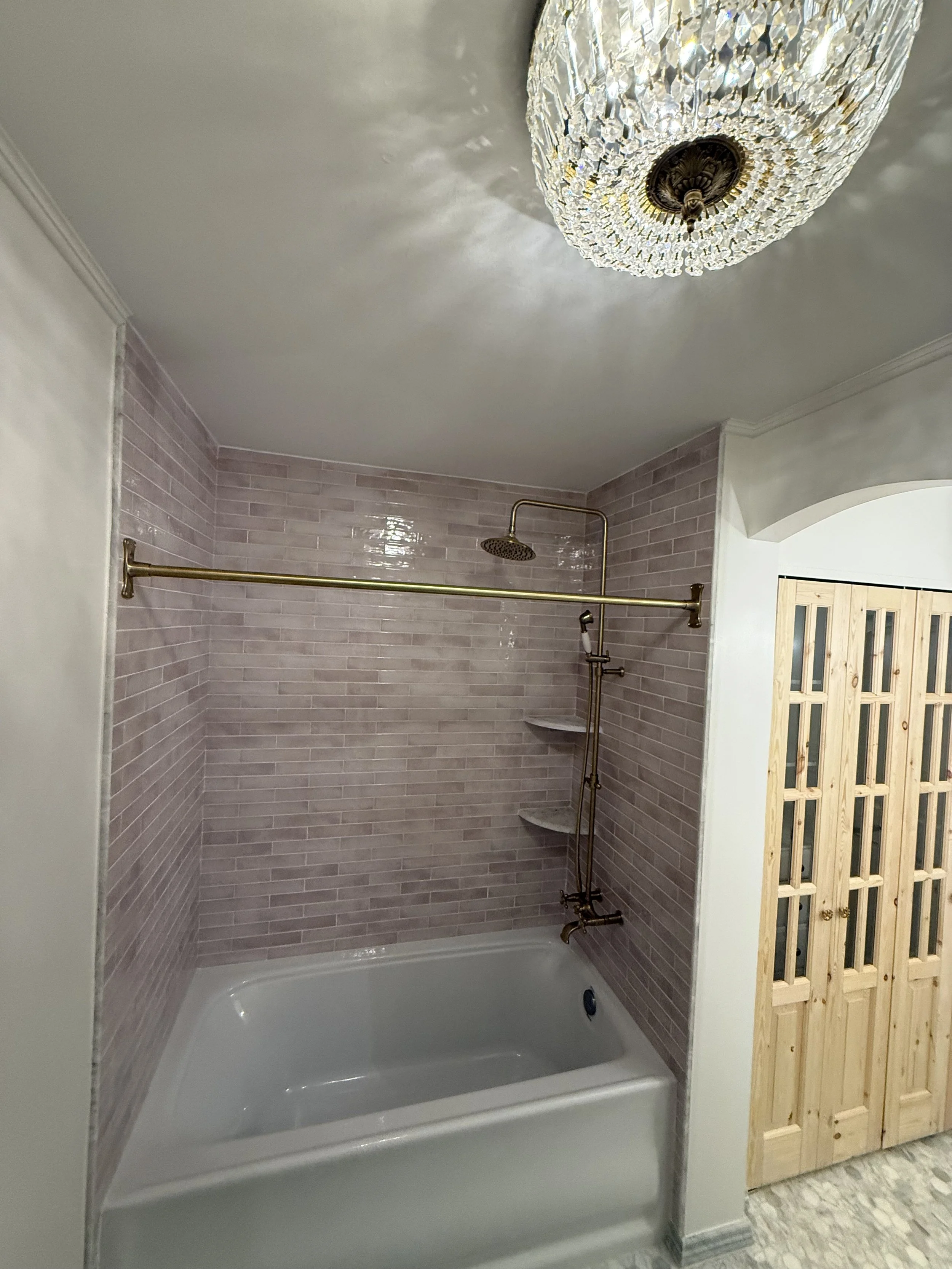 Modern bathroom renovation with a crystal-style ceiling light, gray wall tile, white bathtub, and wood door.