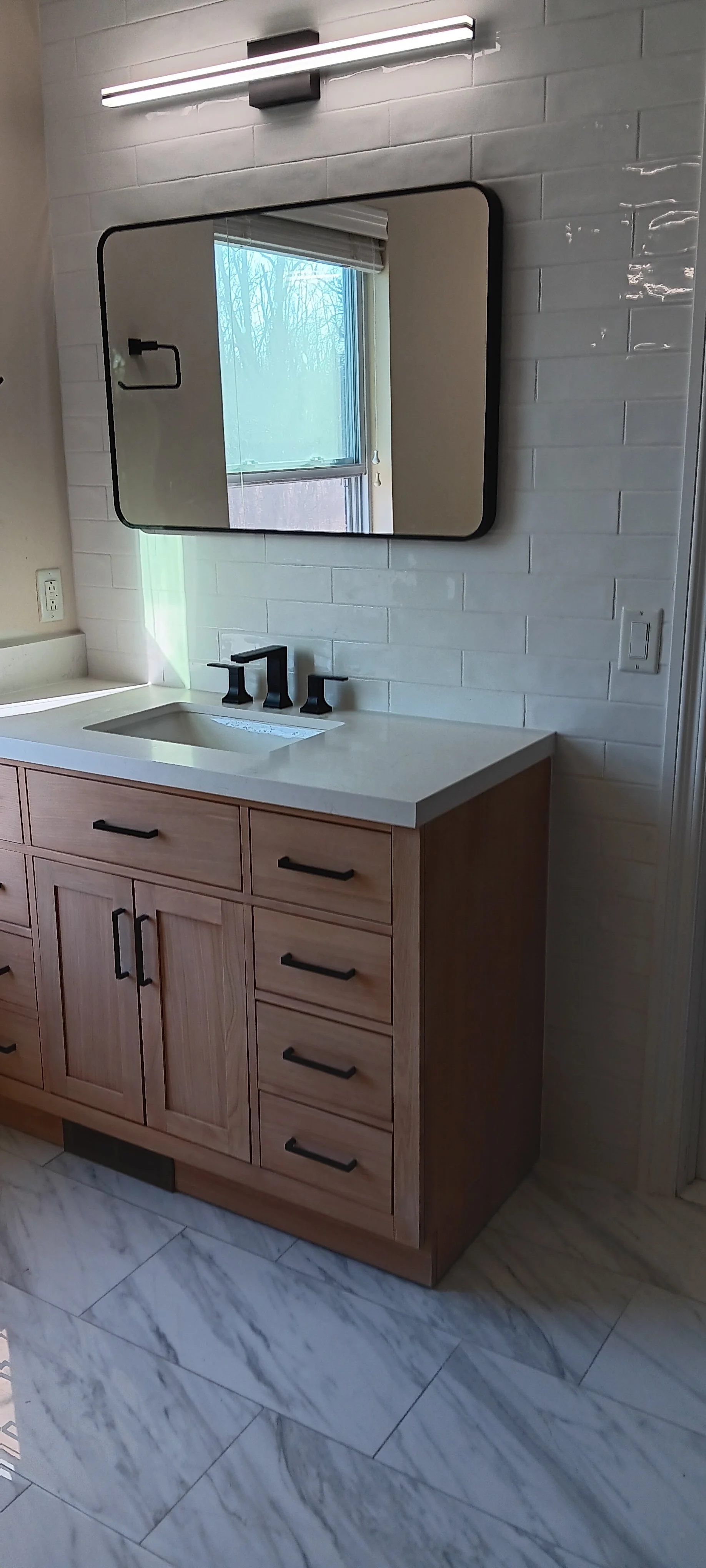 Modern bathroom vanity with wood cabinetry, white countertop, black faucet, framed mirror, and contemporary wall lighting.