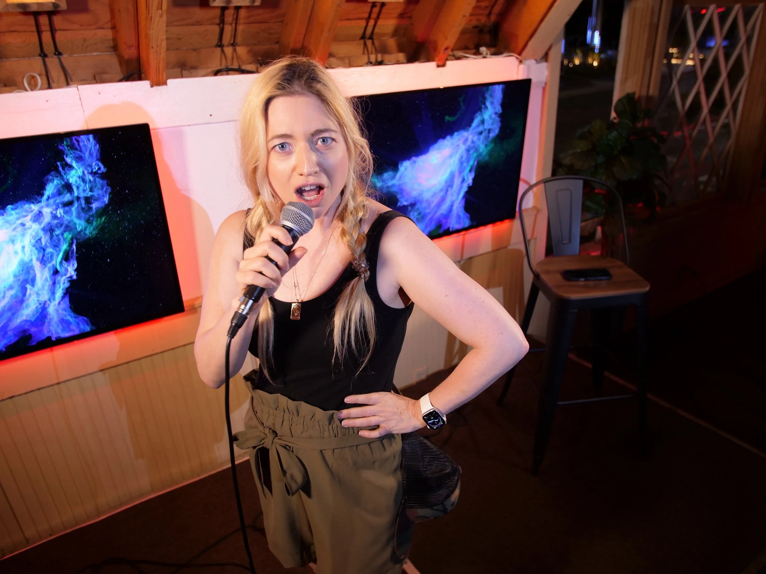 A woman with long blonde hair holding a microphone, standing in front of two large screens displaying colorful cosmic images, in a wooden indoor setting at night.