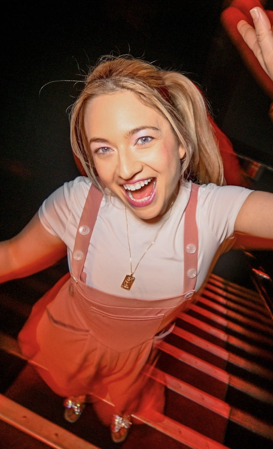 A young woman with blonde hair, smiling and looking up at the camera, stands on a staircase with black and red striped steps. She wears a white shirt, pink overalls, and a necklace, with her arms raised in a joyful pose.