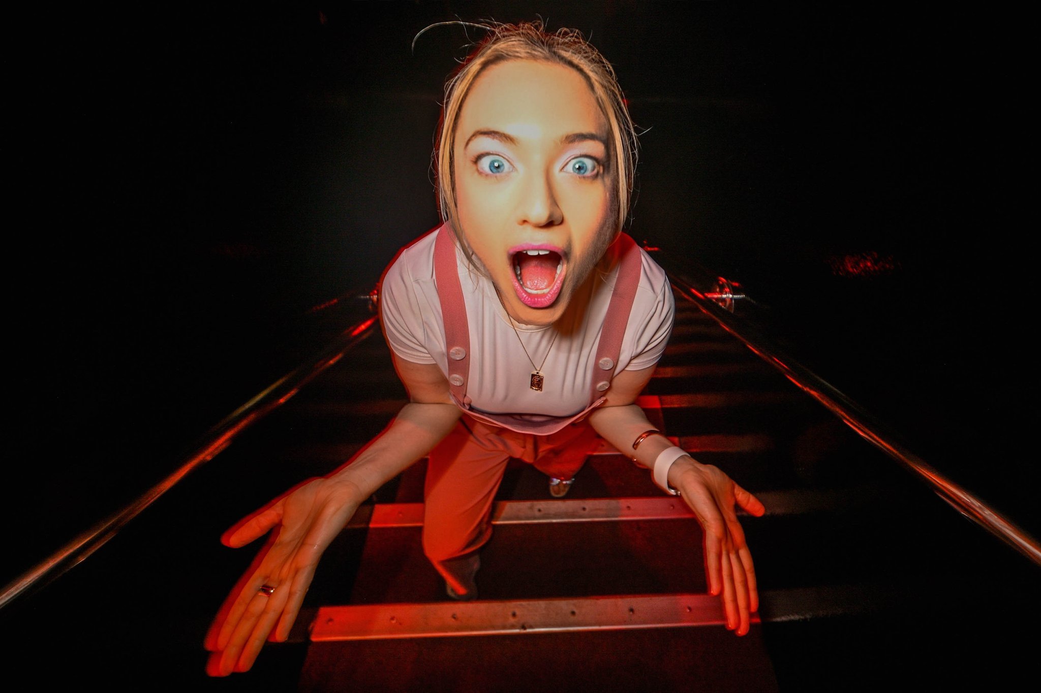 A young woman with blonde hair and blue eyes is laying on train tracks at night with her mouth open in surprise or excitement. She is wearing a pink and white striped shirt and pink suspenders, with streetlights reflecting off the rails.