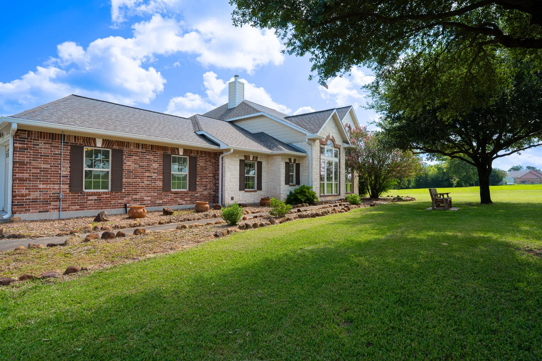 Front view of a brick and stone house with a well-maintained lawn, large trees, and a bright blue sky with scattered clouds.