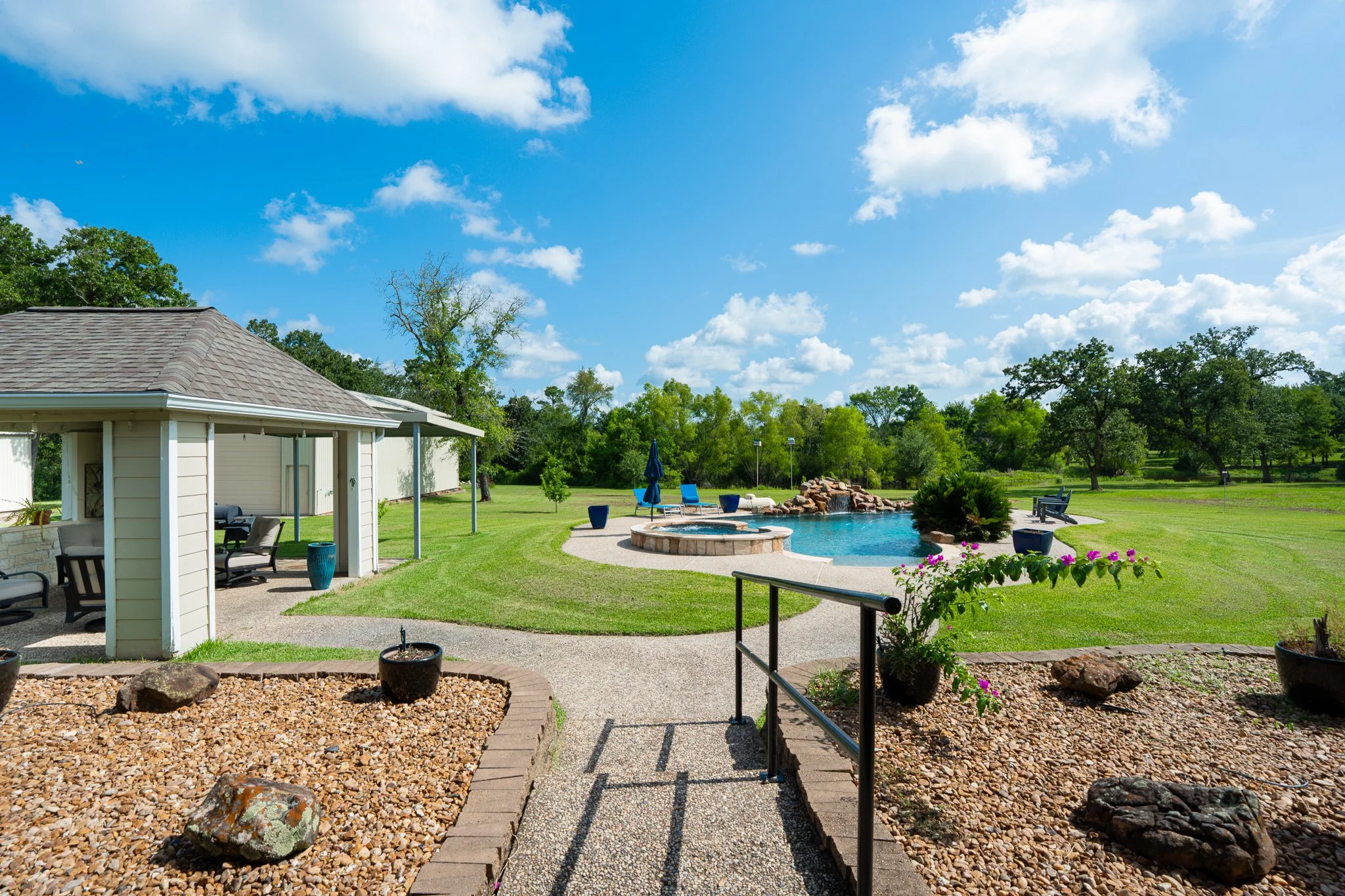 A backyard with a covered patio, a swimming pool, lounge chairs, a hot tub, and lawn with trees under a blue sky with white clouds.