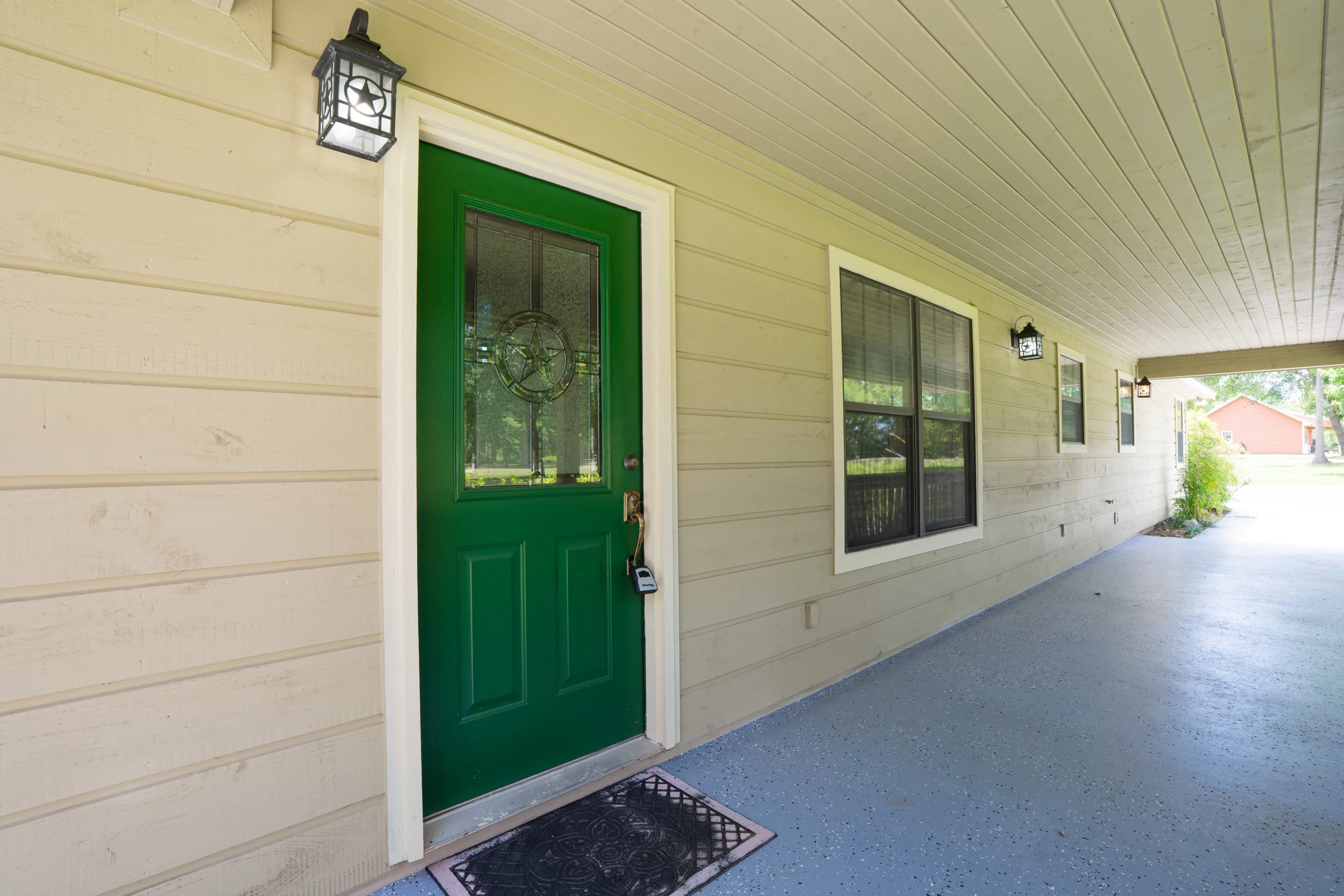 View of a house porch with a green front door with stained glass, beige siding, two black outdoor wall lanterns, and a striped doormat.