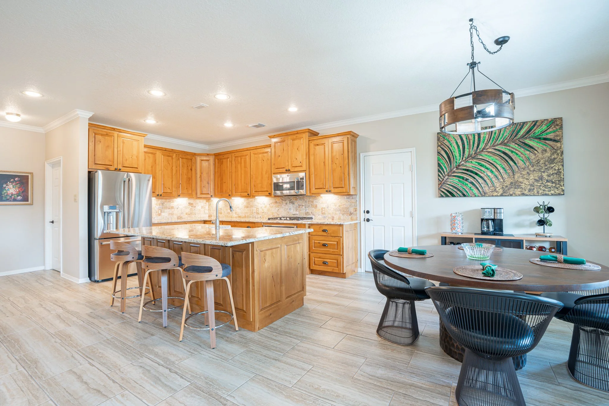 A modern kitchen and dining area with wooden cabinets, a granite island, stainless steel appliances, a round dining table with black wire chairs, and decorative wall art featuring green leaves.