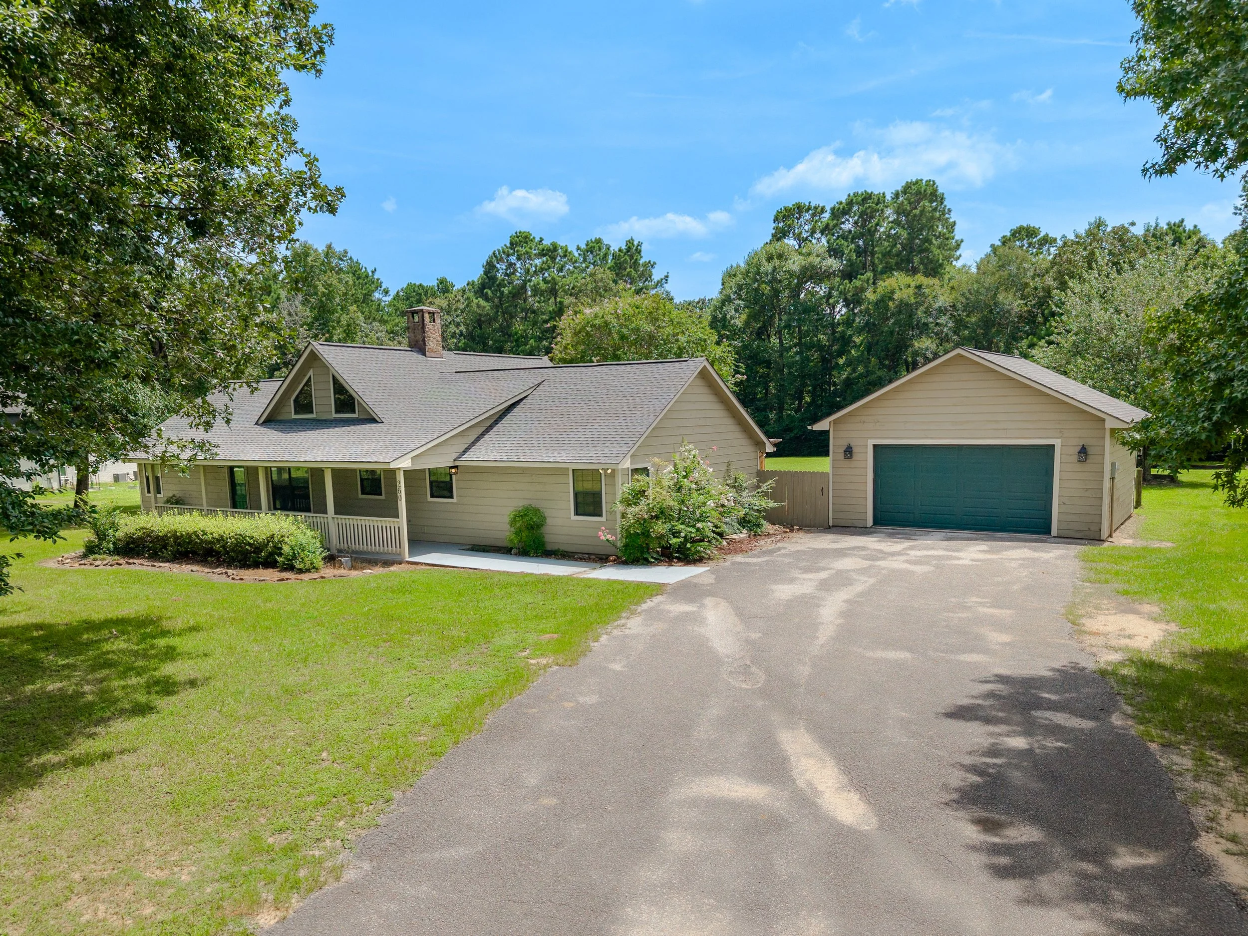 A suburban house with beige siding, a dark-colored garage door, and a front porch, surrounded by green grass, trees, and a driveway on a sunny day with a blue sky and some scattered clouds.