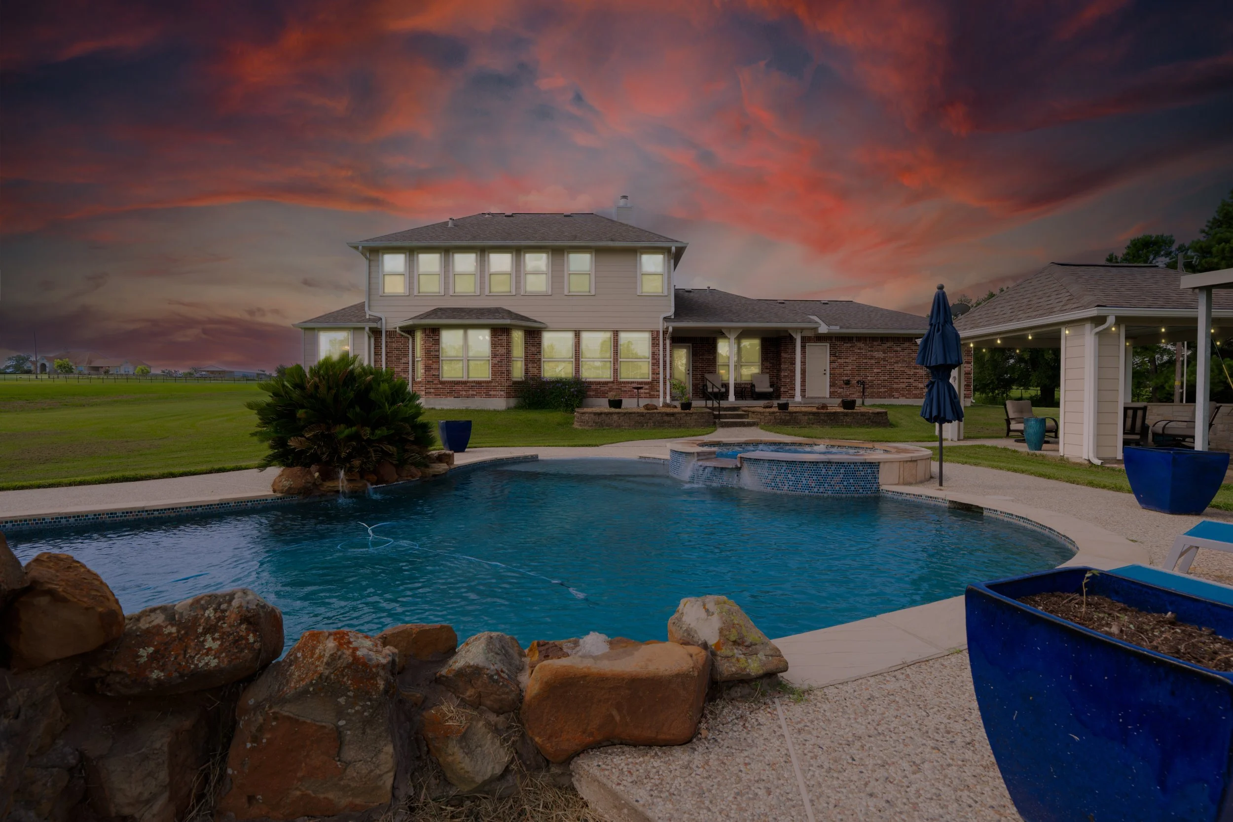 A backyard featuring a swimming pool with rocks at the edge, a covered patio area with outdoor furniture and an umbrella, well-maintained grass, and a large house with multiple windows and a brick and siding exterior, against a vibrant sunset sky with colorful clouds.